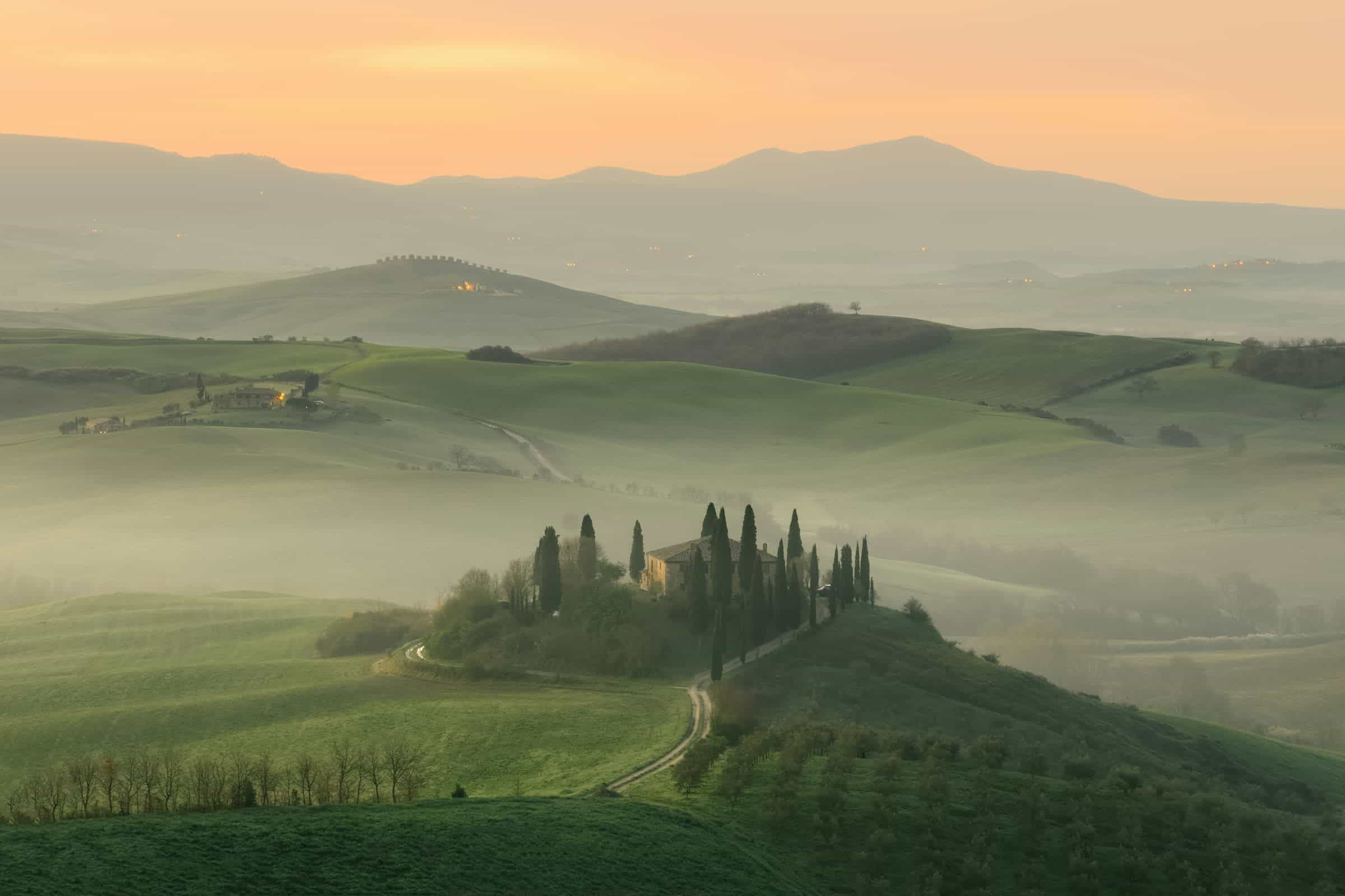 Aerial landscape of a misty morning with the sun rising over the hills in the distance and cypress trees surrounding a country house