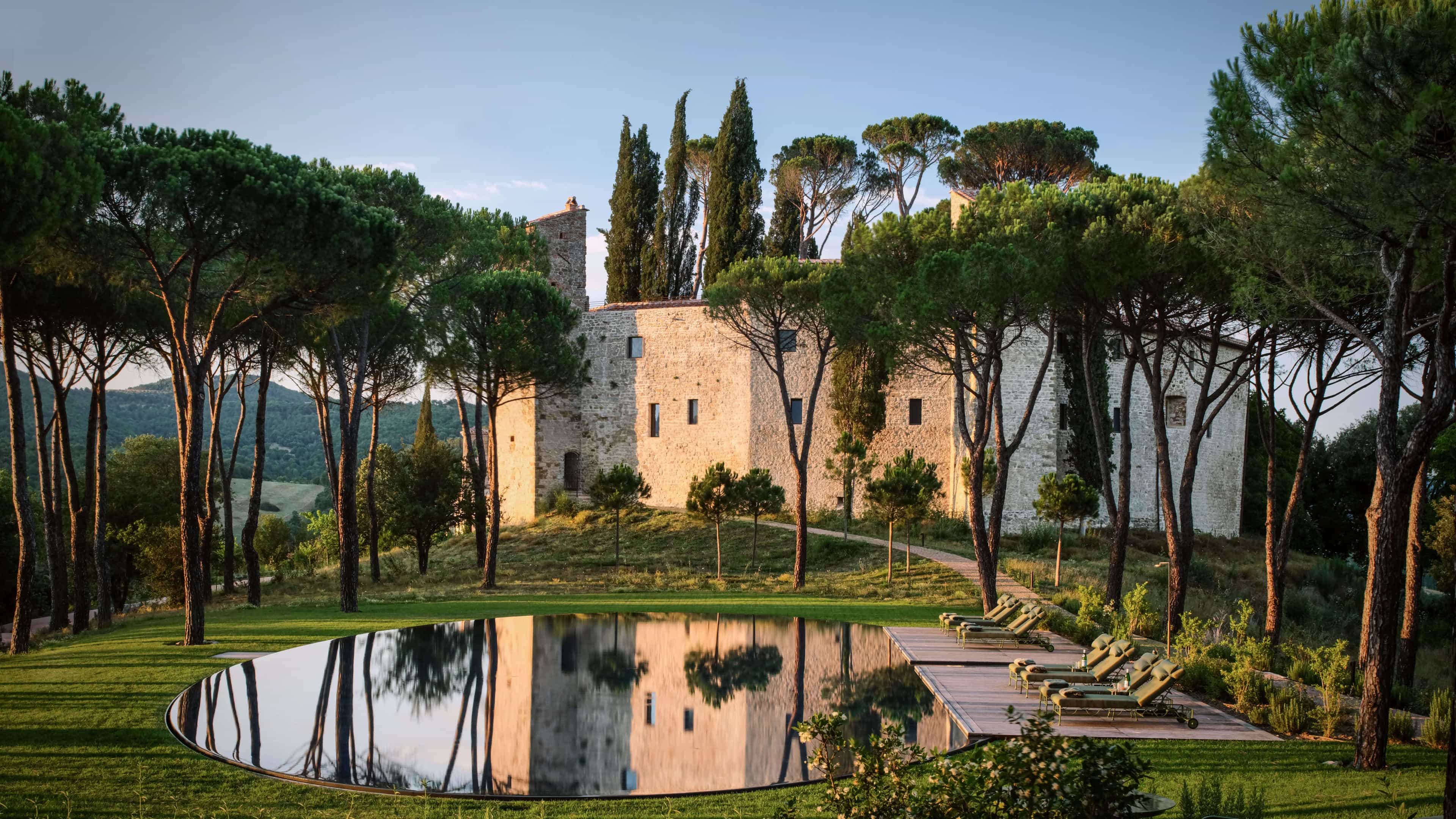 A pool surrounded by pine trees and ancient tan stone building