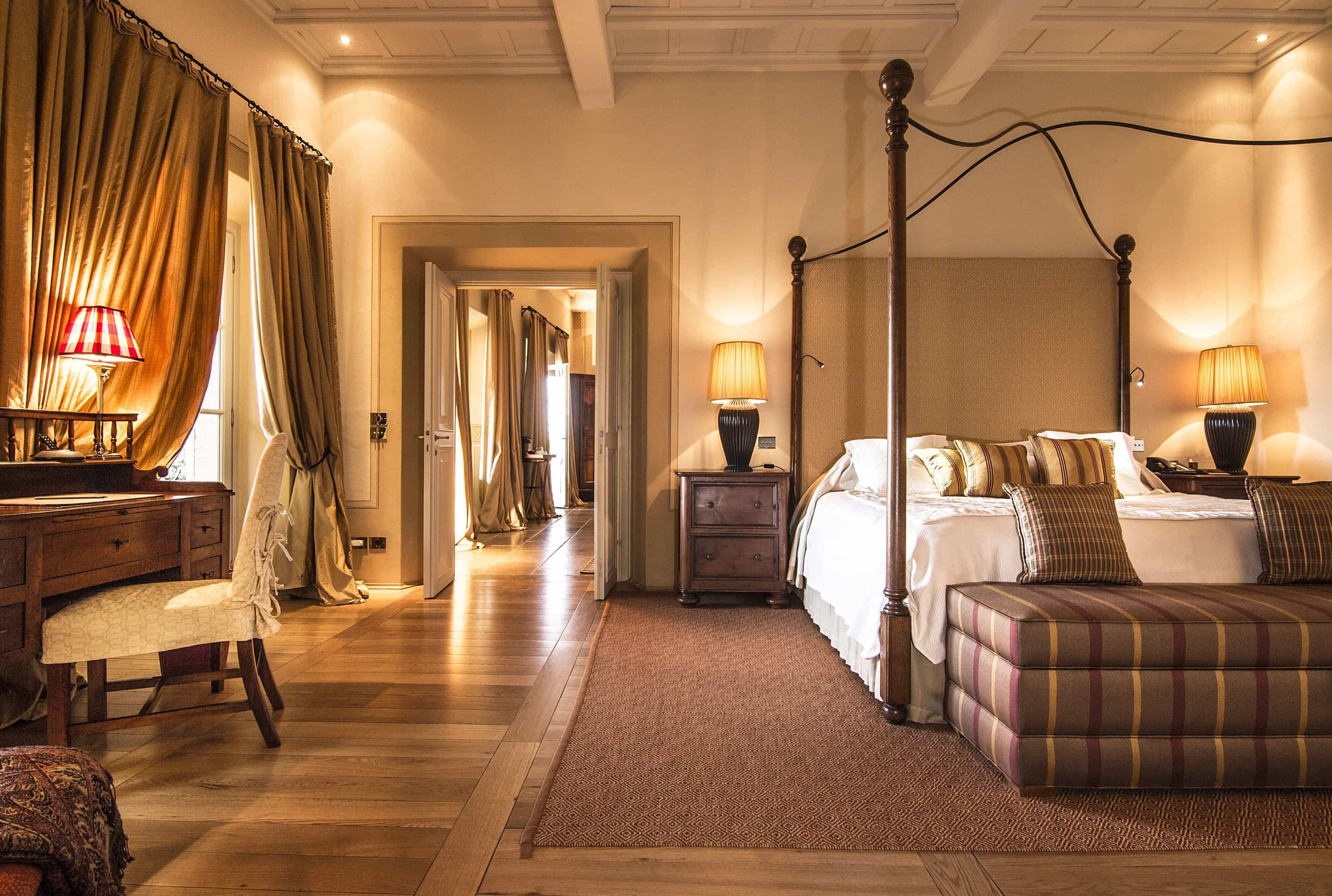 A four poster metal bed with white wood beam ceiling, dark wood floor and desk with chair off to the side