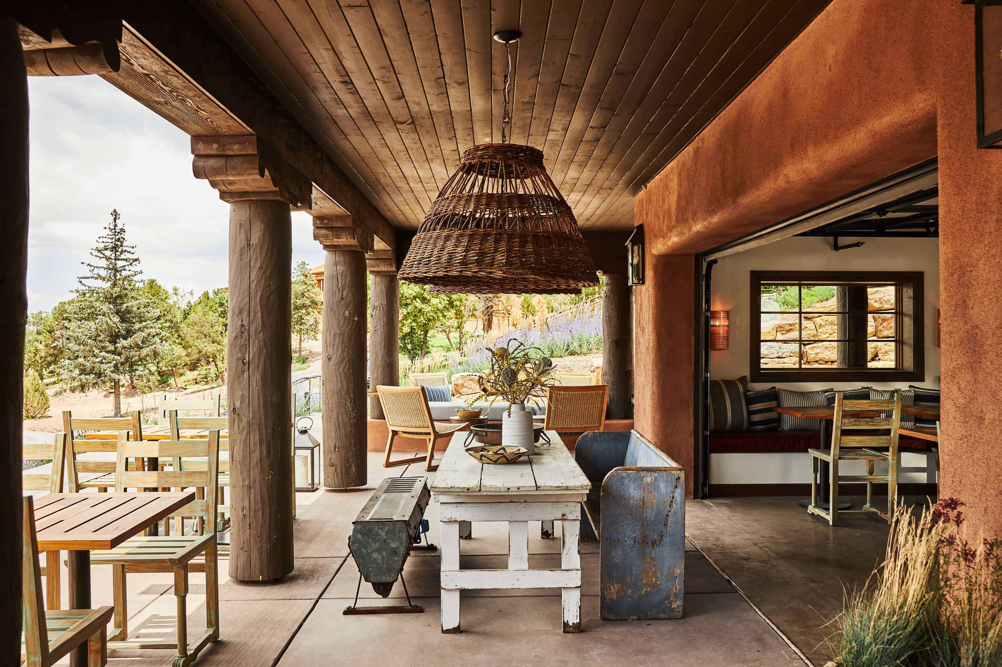 Rustic outdoor dining area with wooden columns, a large woven pendant light, mismatched furniture, and views of a garden with trees and lavender.