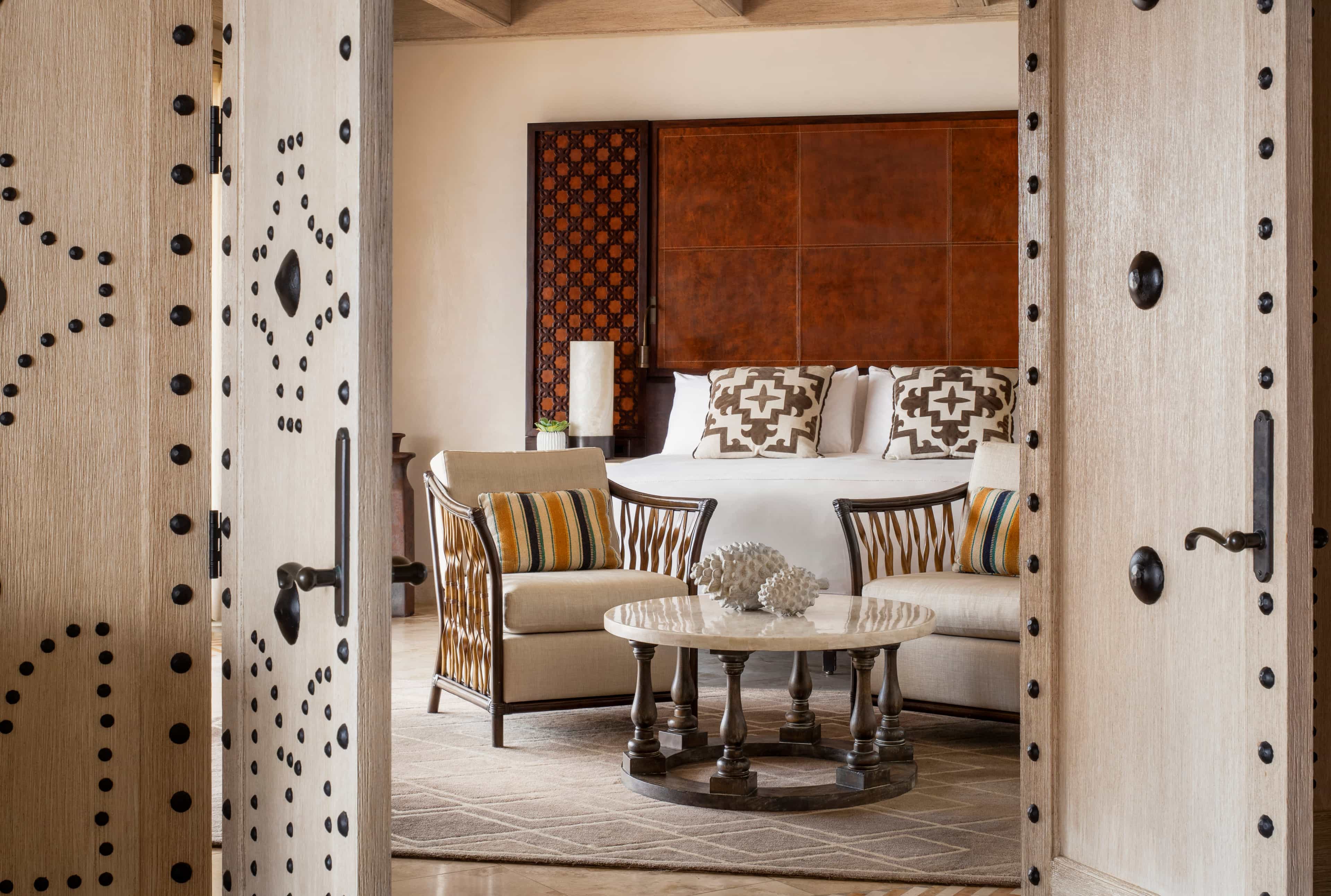 View through ornate wooden double doors into a luxurious hotel suite with a king bed, patterned pillows, upholstered armchairs, and a round coffee table topped with decorative coral.