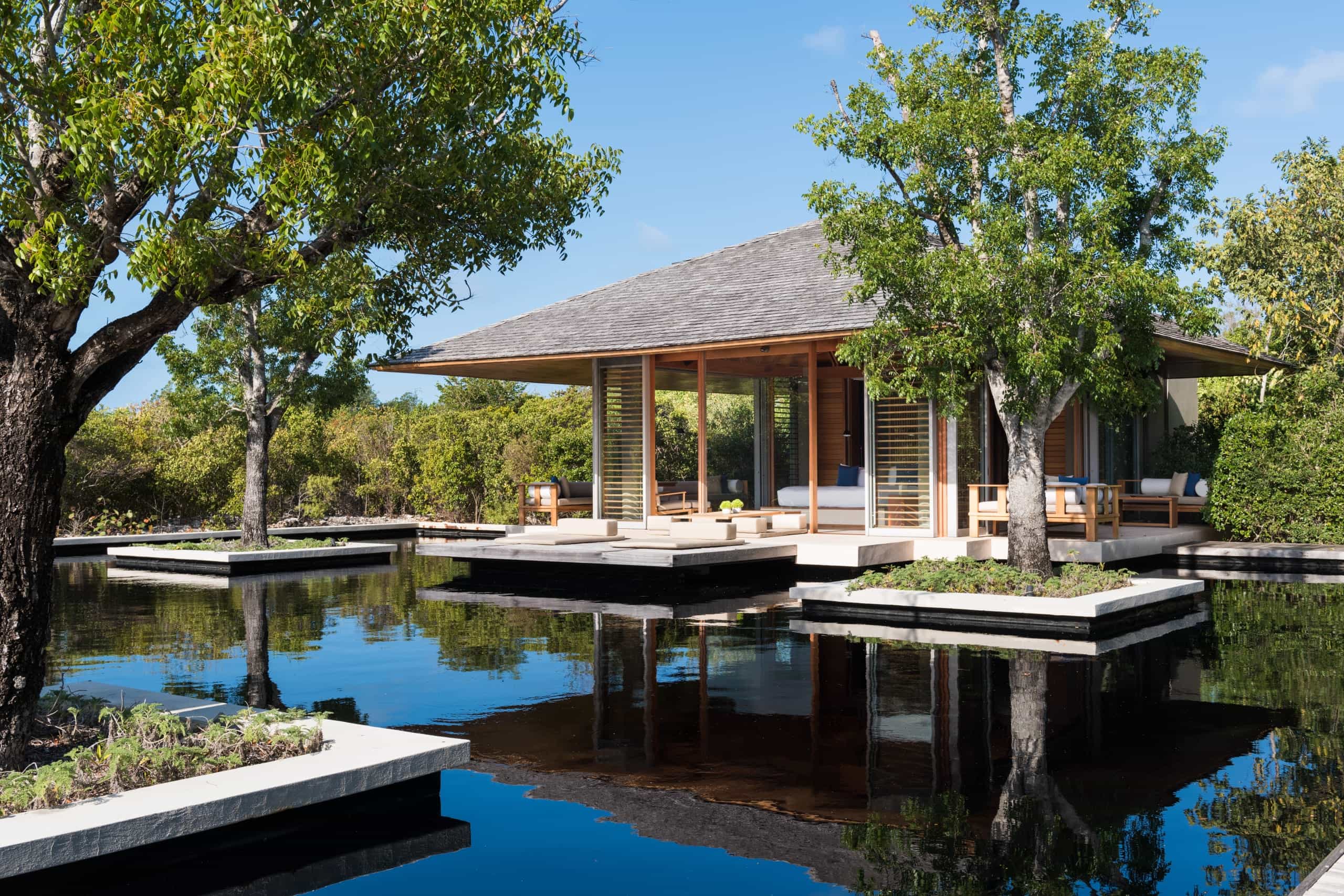 Modern open-air pavilion with wood and glass design, surrounded by tranquil reflecting pools and lush greenery, with trees growing from raised planters in the water.