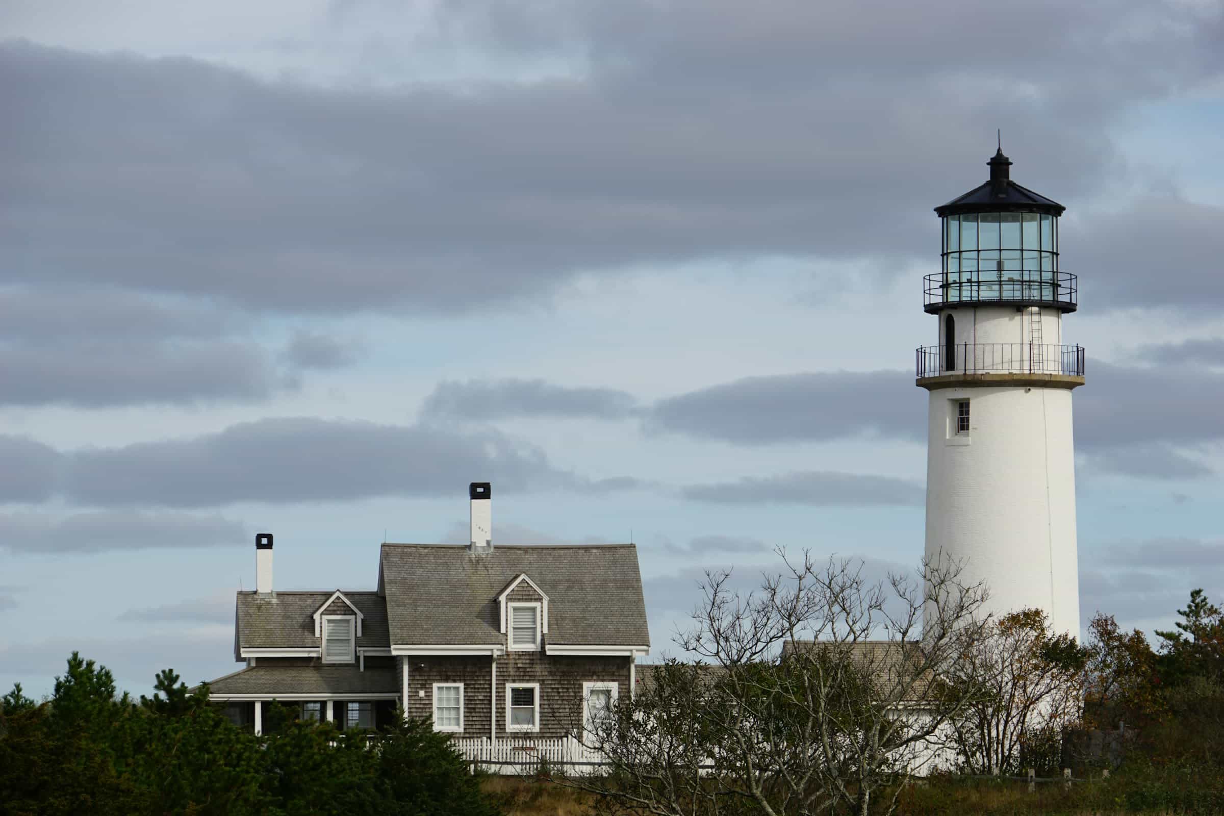 a white lighthouse and a grey shingle house on a cloudy day