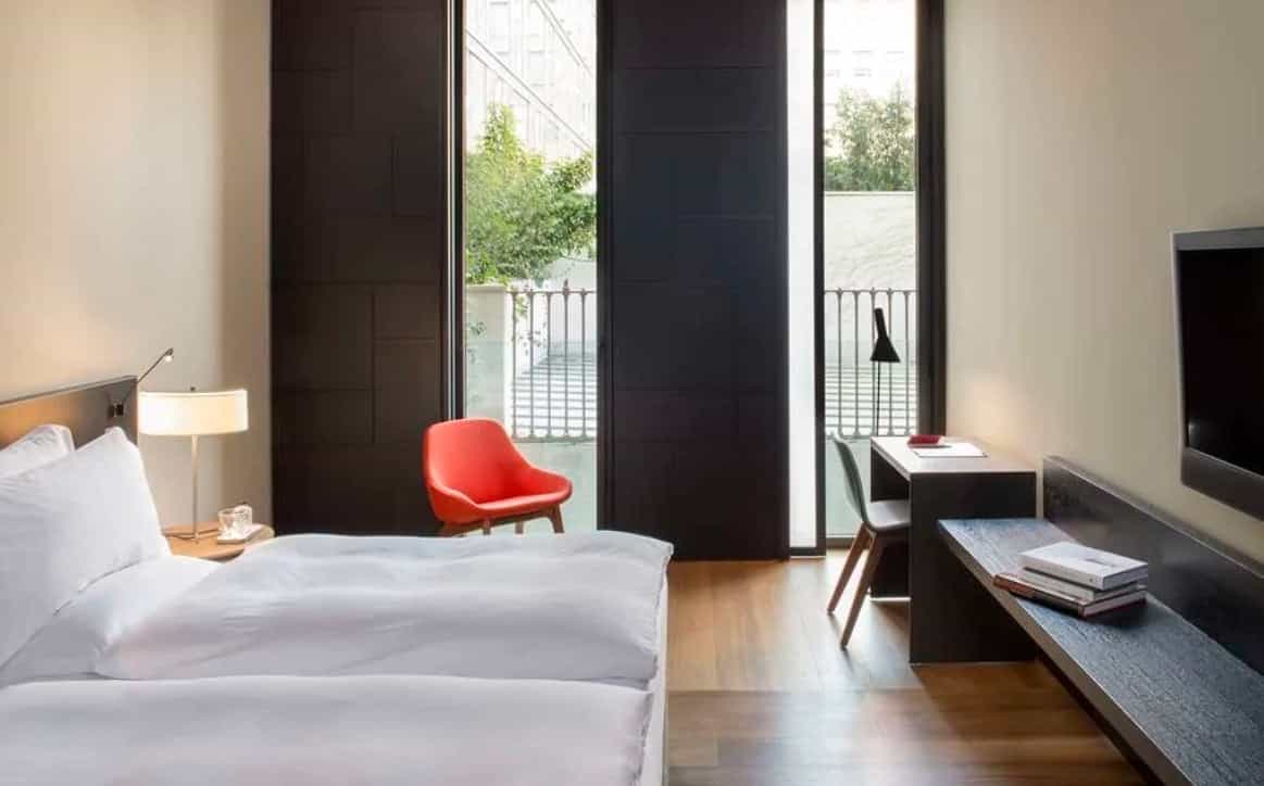 Contemporary hotel room with neutral colors and dark window shutters, a desk and desk lamp, and bright red armchair.