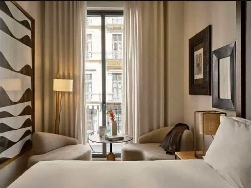 An elegant, modern hotel room with neutral furnishings and wall artwork with tall window and city views.