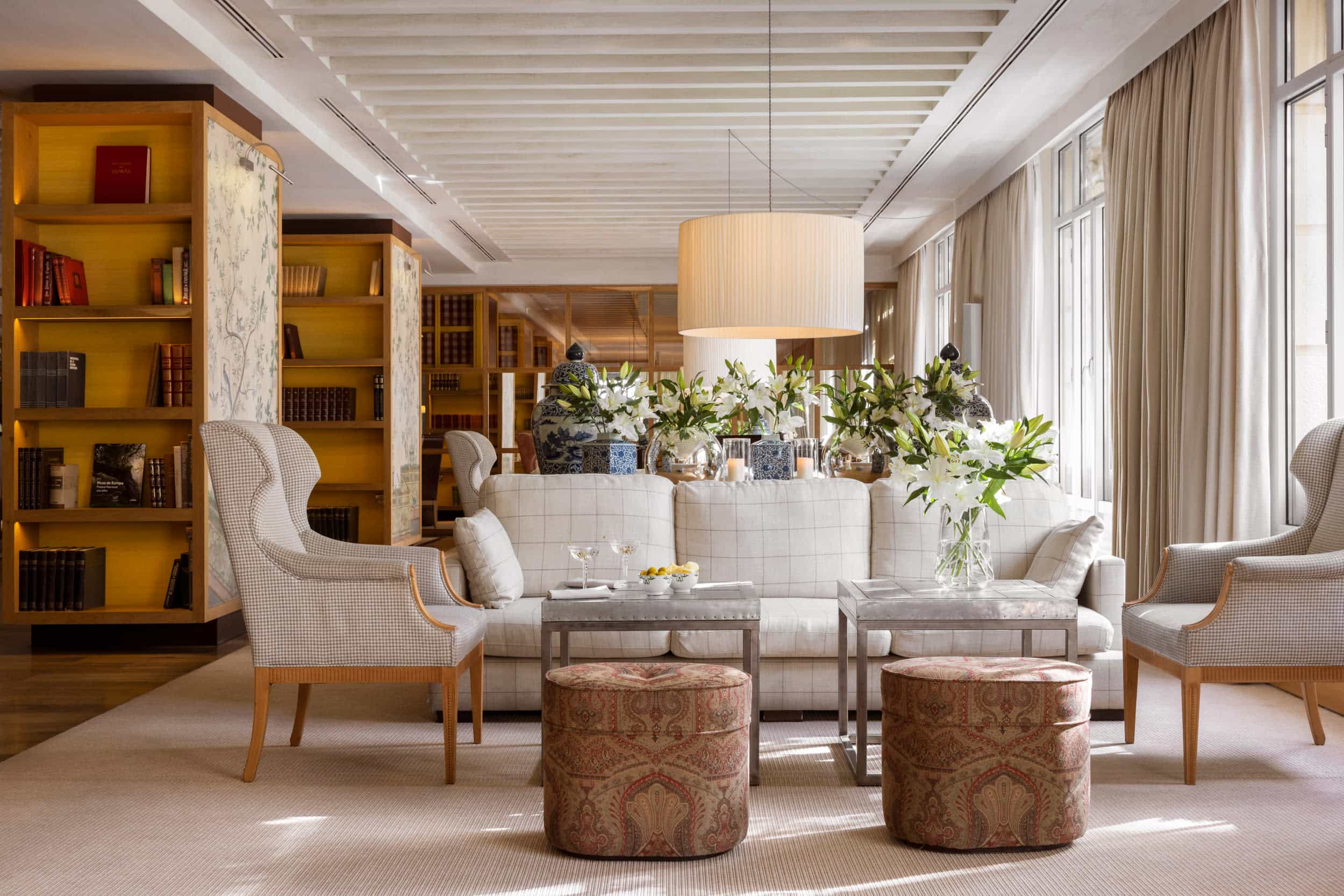 An elegant hotel lobby area with neutral decor and furnishings with wooden accents, bookshelves and floor-to-ceiling windows with drapes.