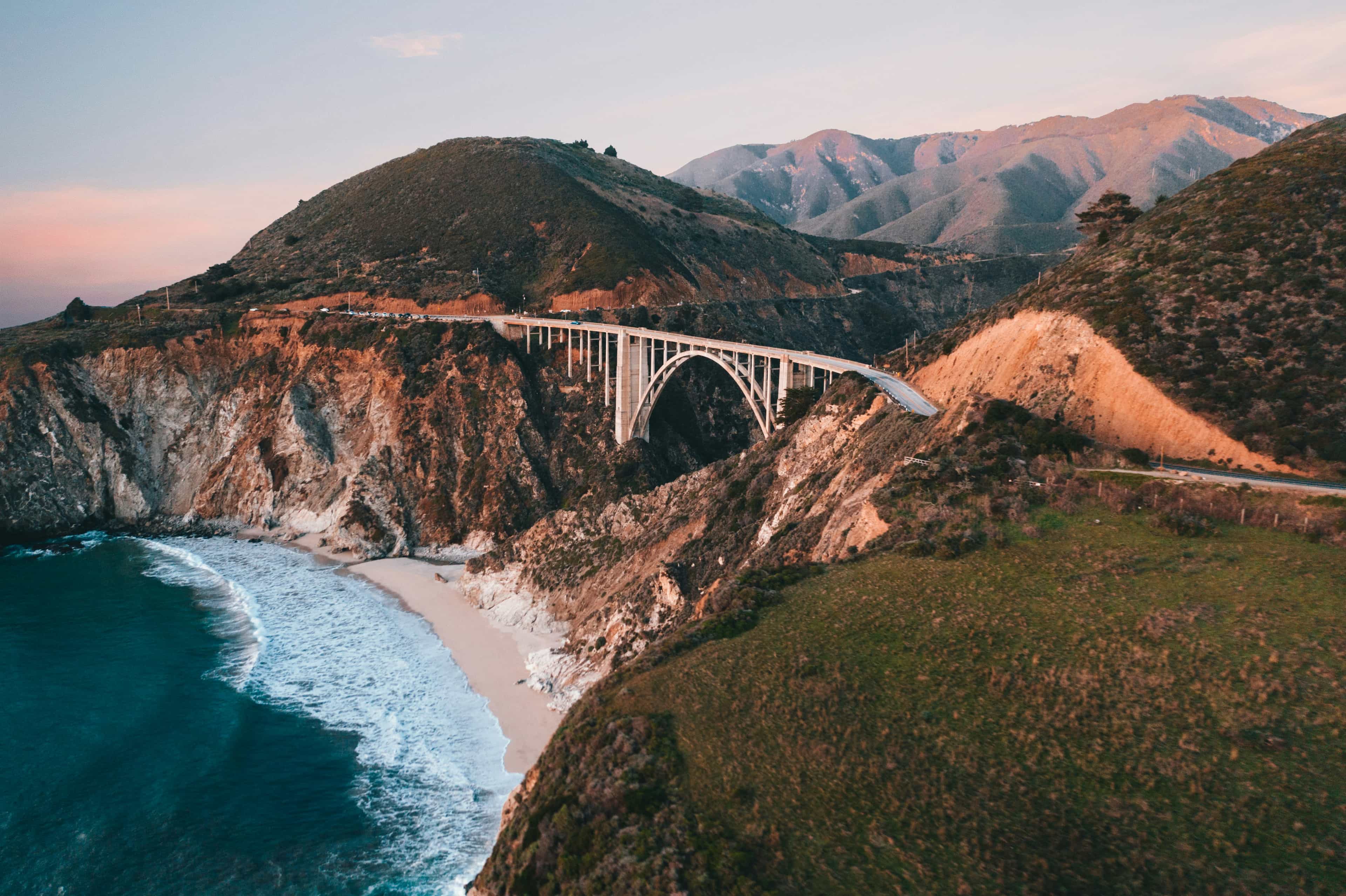 A stunning coastal bridge arching over rugged cliffs beside the Pacific Ocean, surrounded by rolling hills and a winding coastal road during golden hour.