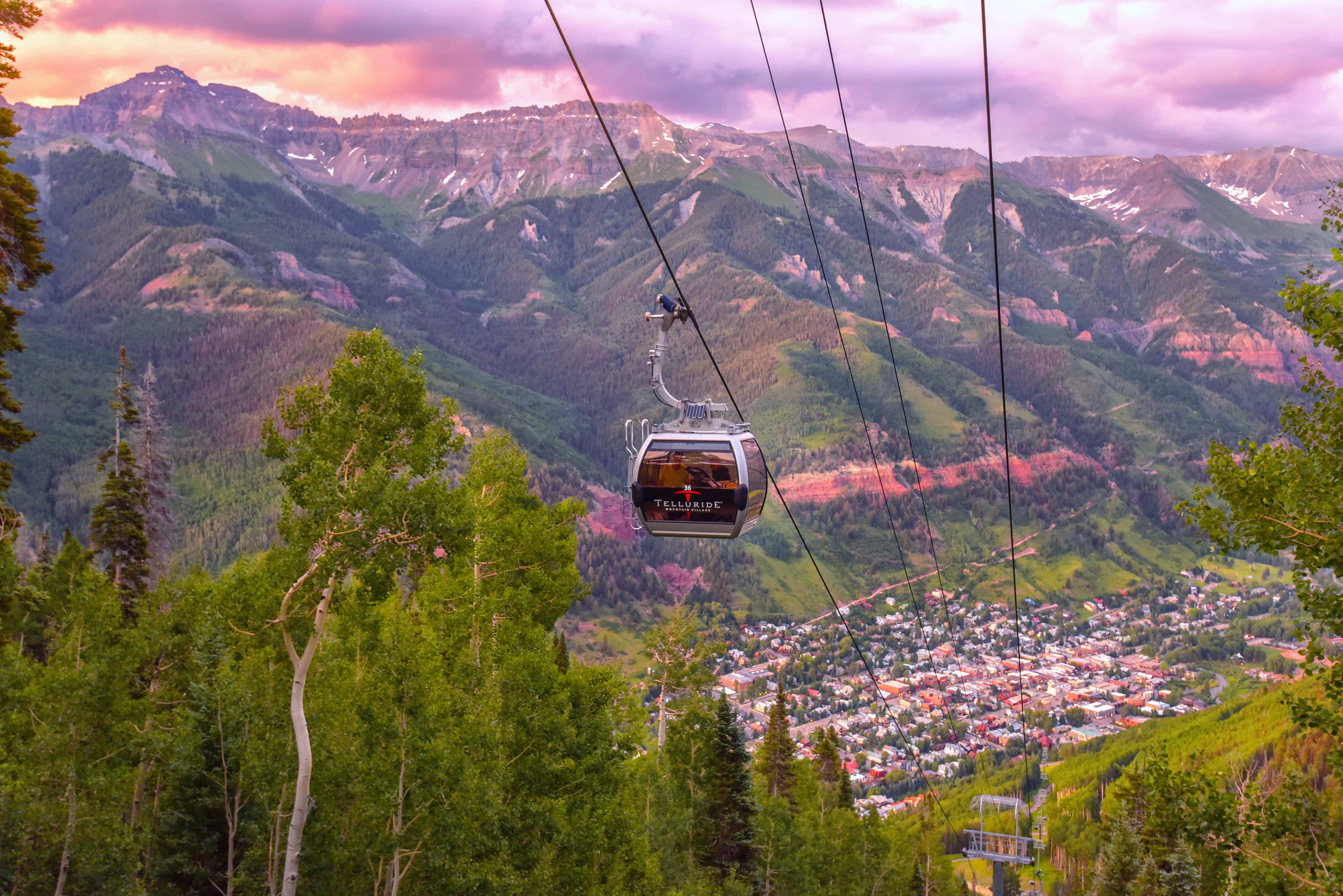 A gondola lift cabin suspended on cables above a lush green forest with a colorful mountain range and a small town in the valley below during sunset.