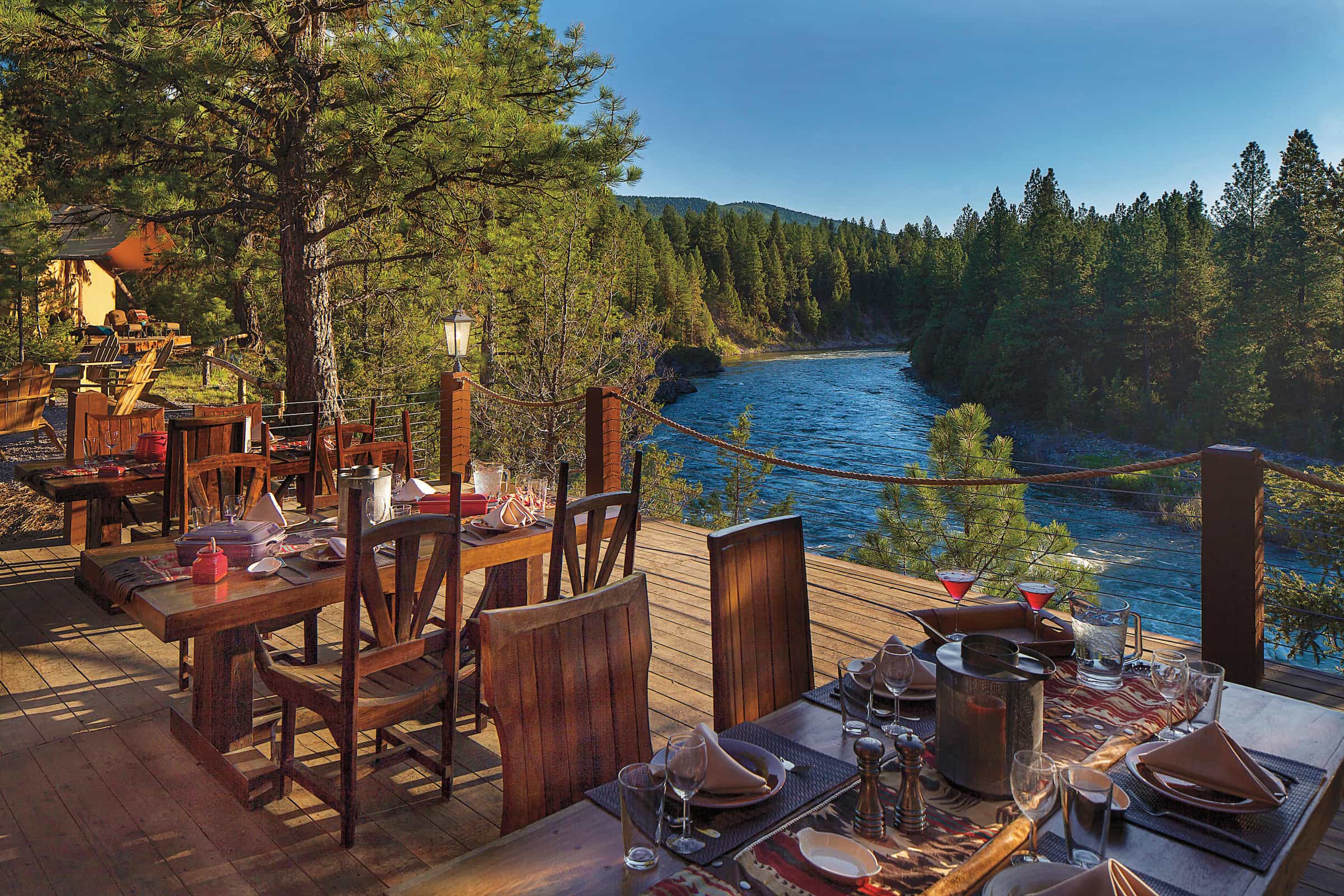 Outdoor dining area on a wooden deck overlooking a river and forest, with tables set for a meal and surrounded by pine trees under a clear blue sky.