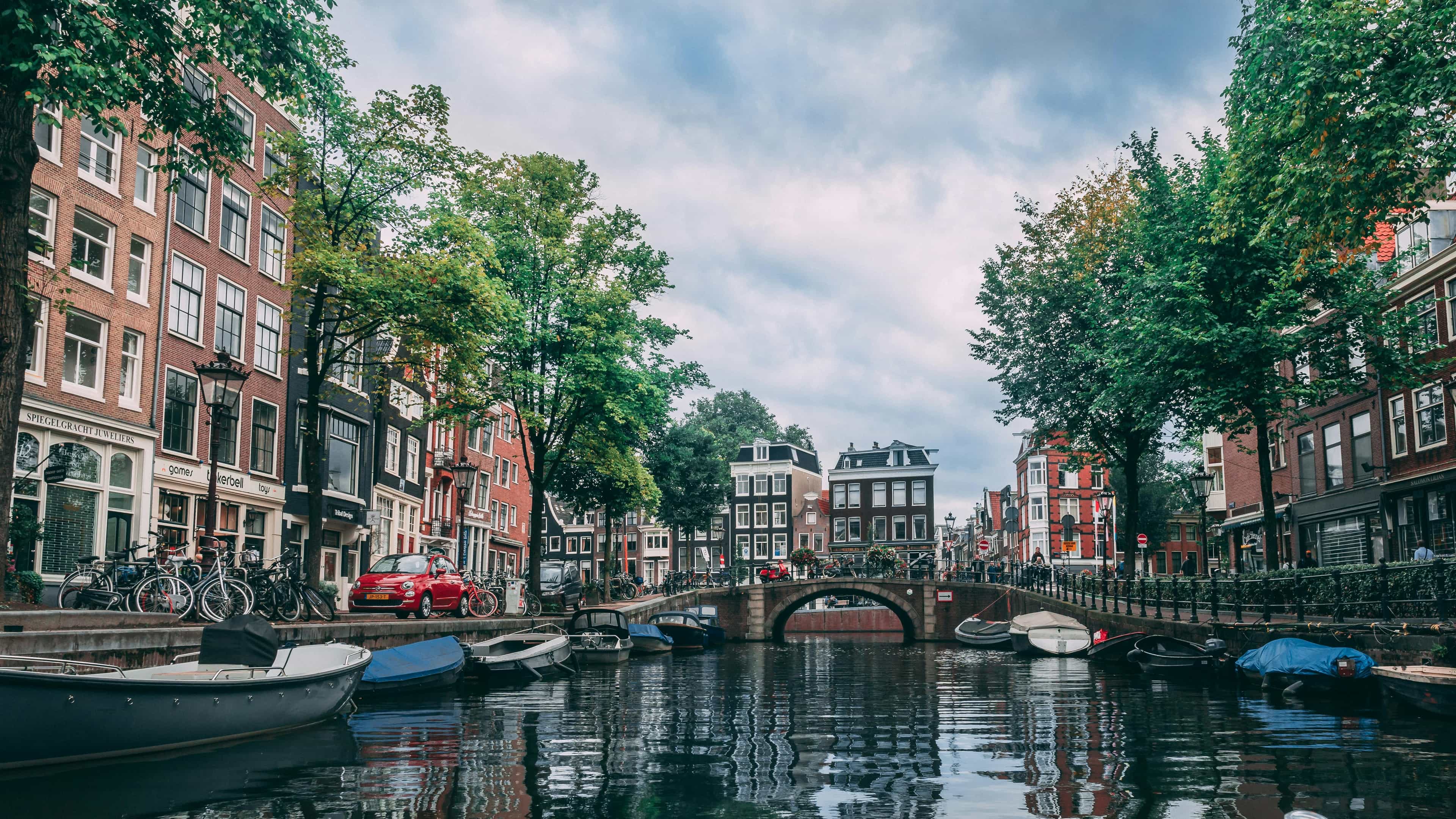 boats lining a calm canal with brick buildings and trees along them during day
