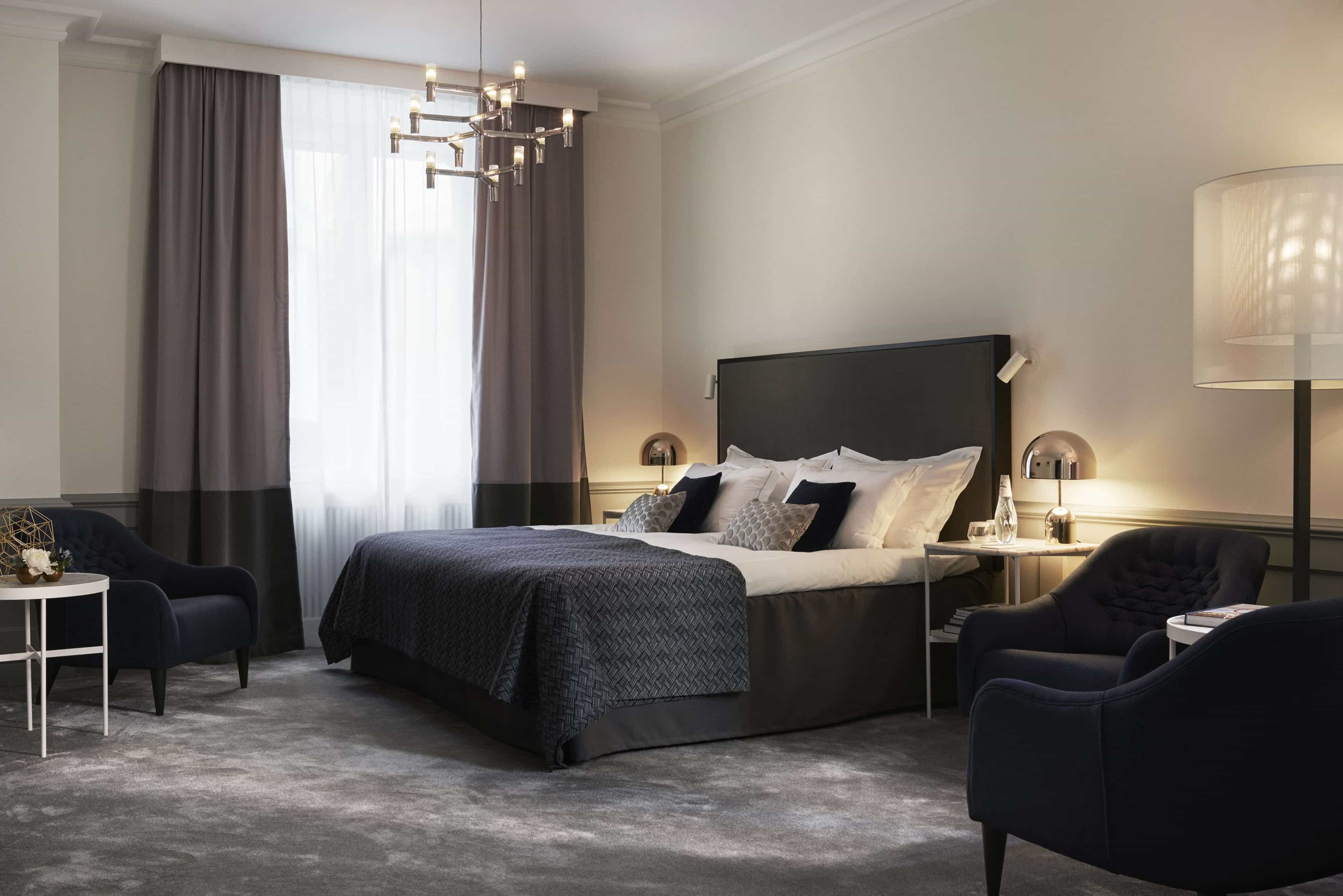 Modern hotel room with dark a blue bed and chair, grey carpet, modern chandelier, and white walls