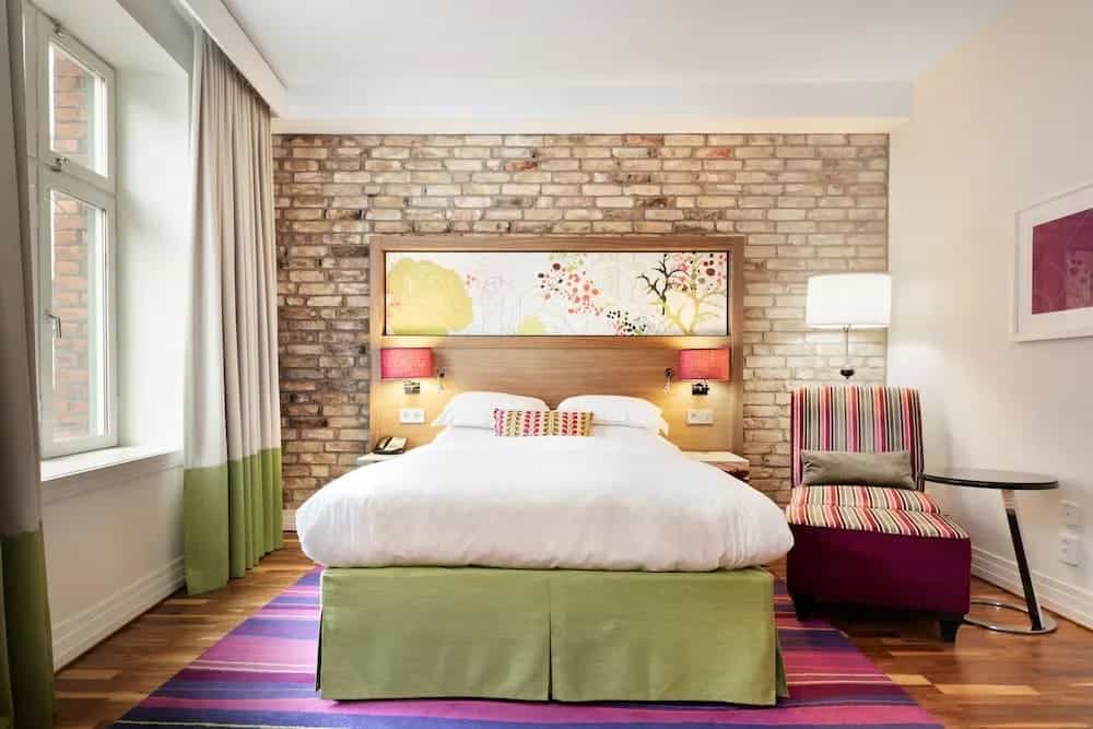 bright hotel room with multi colored purple and pink carpet, green bed skirt, and floral headboard with brick wall.