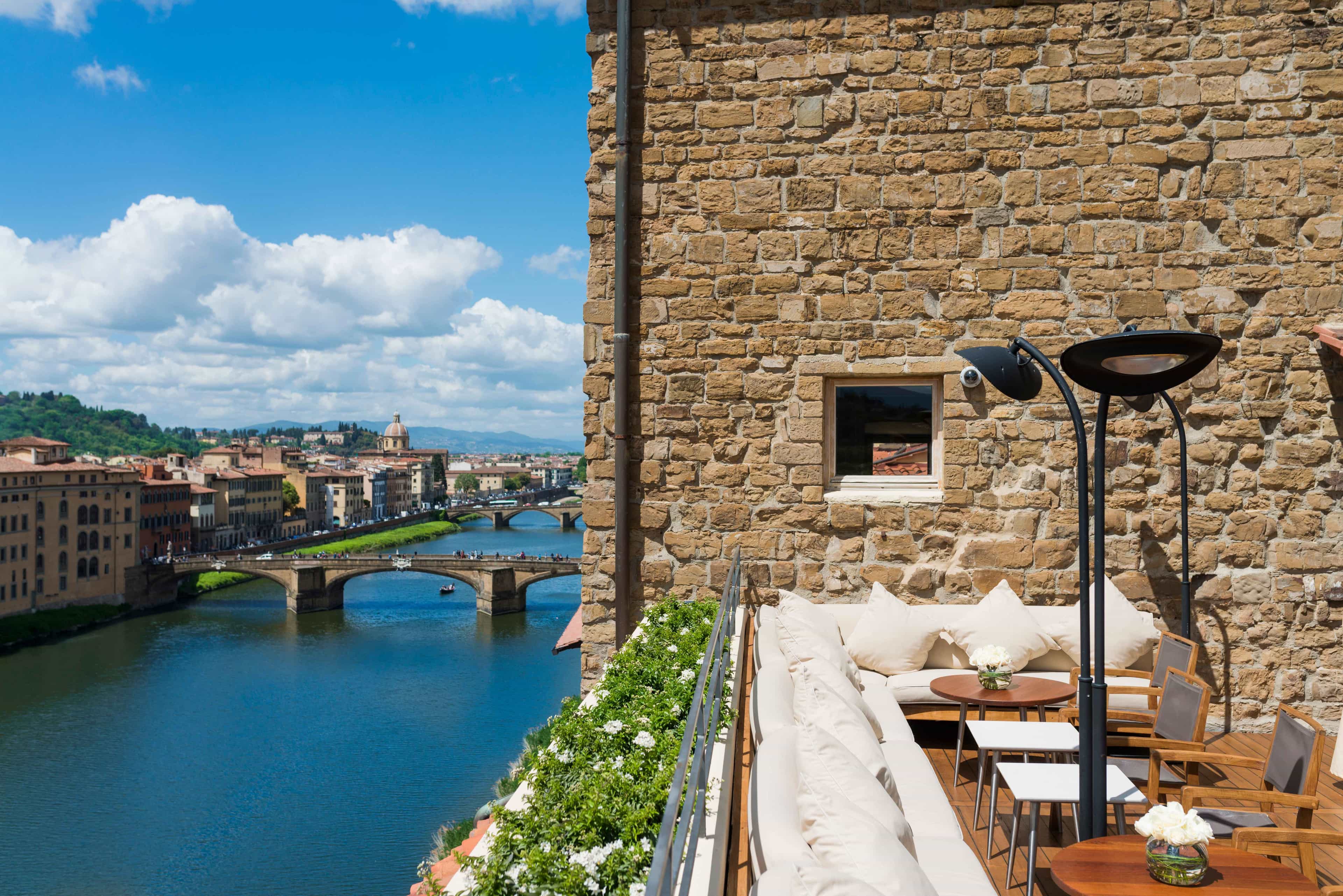 A view of a rooftop terrace with cream lounge seating around the edge against a stone wall and overlooking a river and bridge