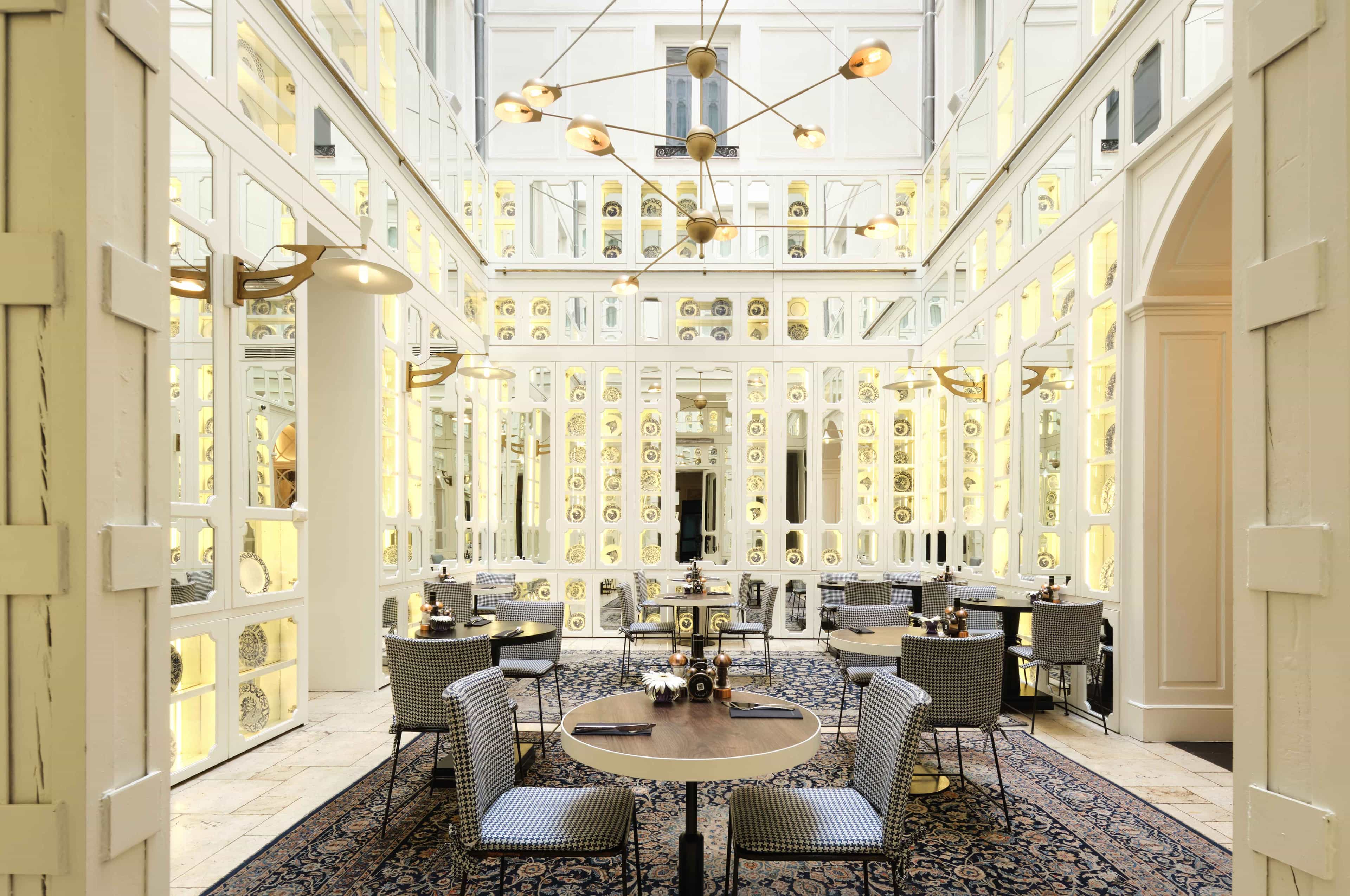 Bright restaurant with patterned chairs, round tables, and mirrored walls showcasing decorative plates under warm modern lighting.