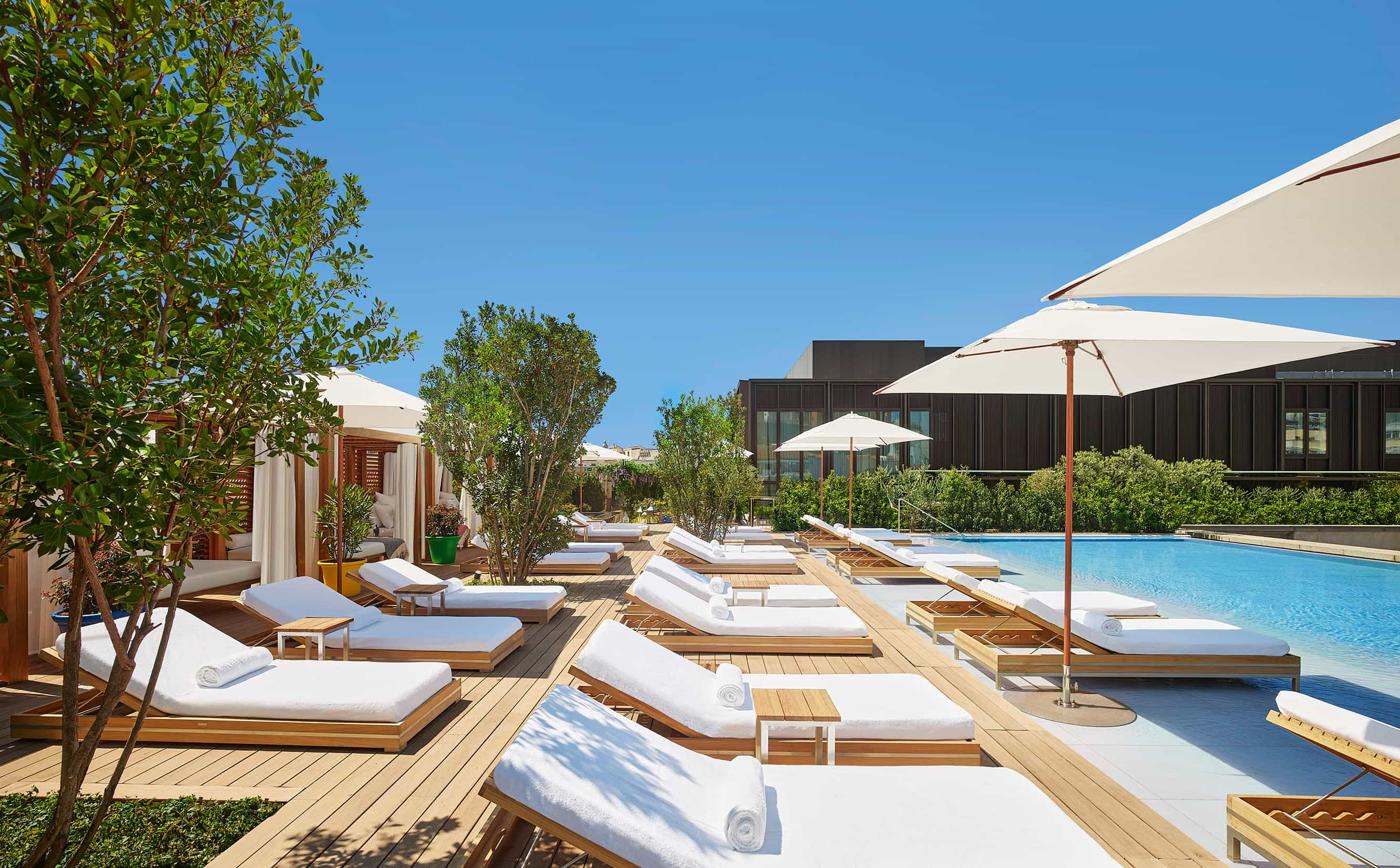 Outdoor pool deck with white sun loungers, umbrellas, and cabanas surrounded by greenery under a clear blue sky.