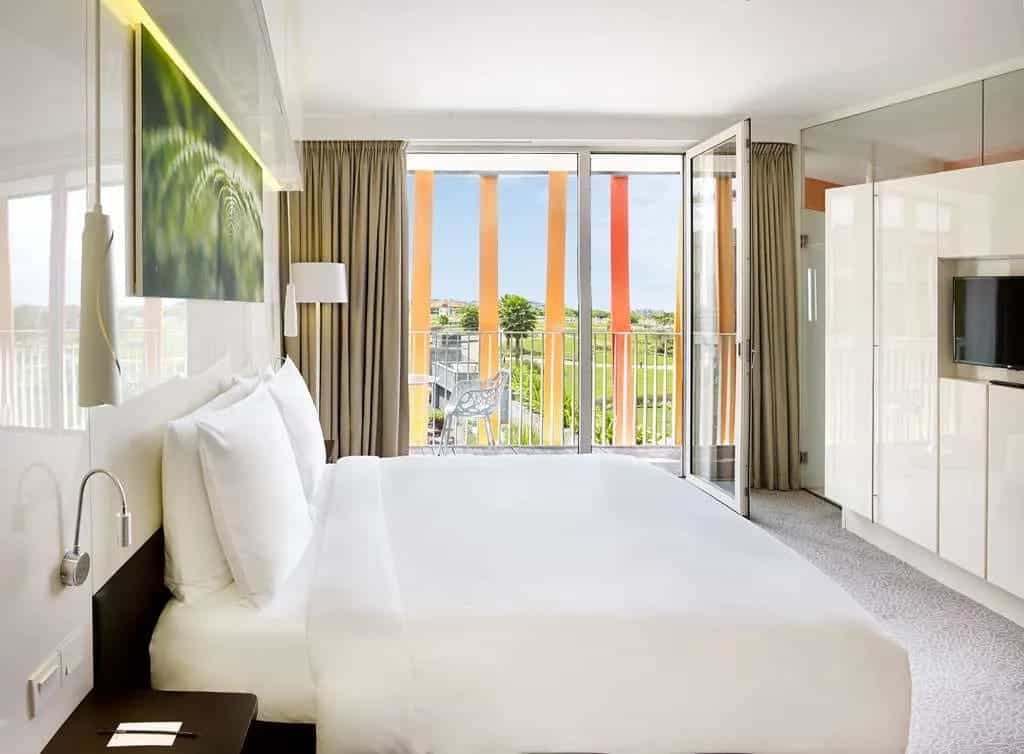 white hotel bed with green landscape photo above bed and windows looking outside with bright orange modern metal siding