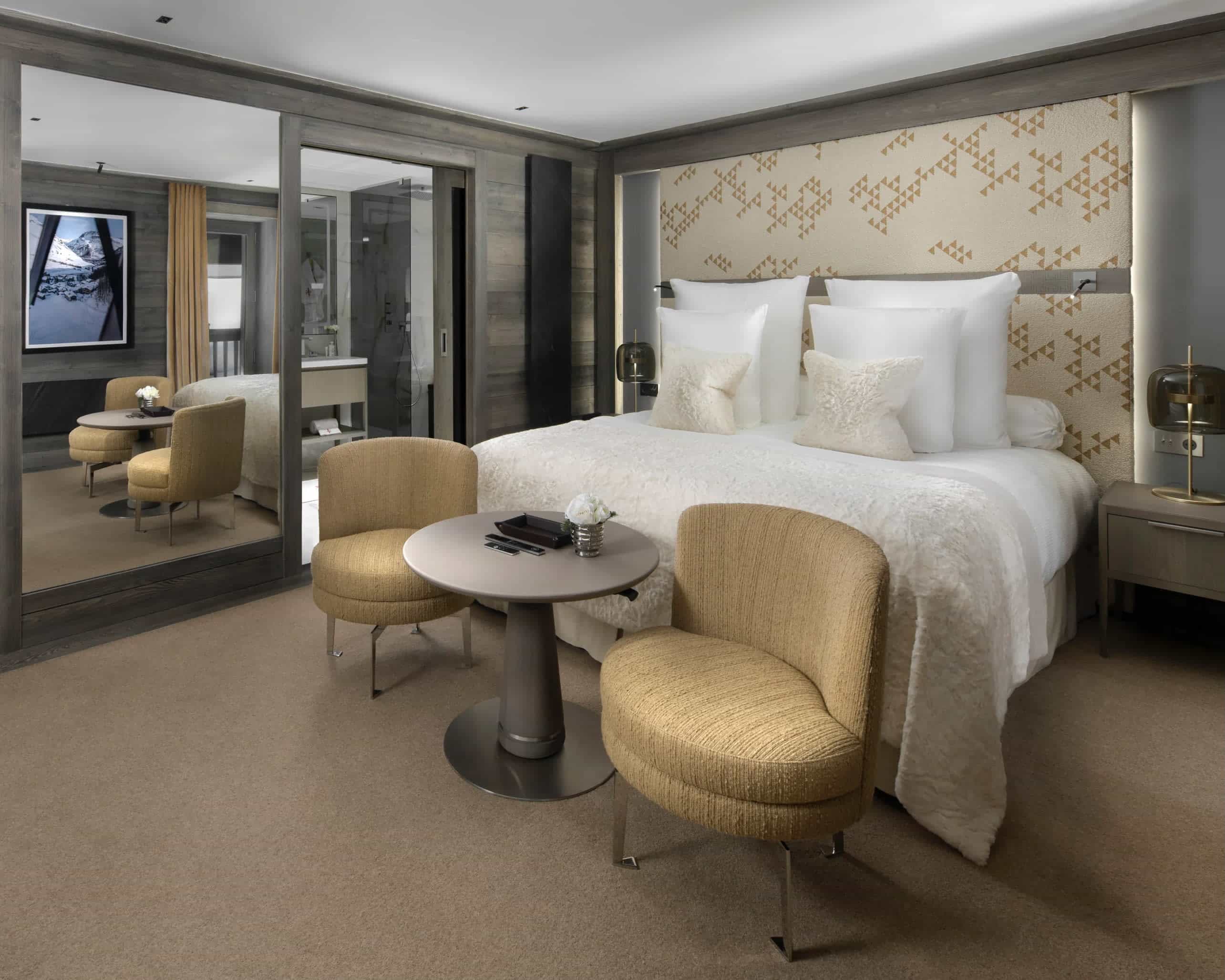 A luxurious modern room with white bed linens, gold chairs at foot of the bed, beige headboard, and glass closet next to the bed