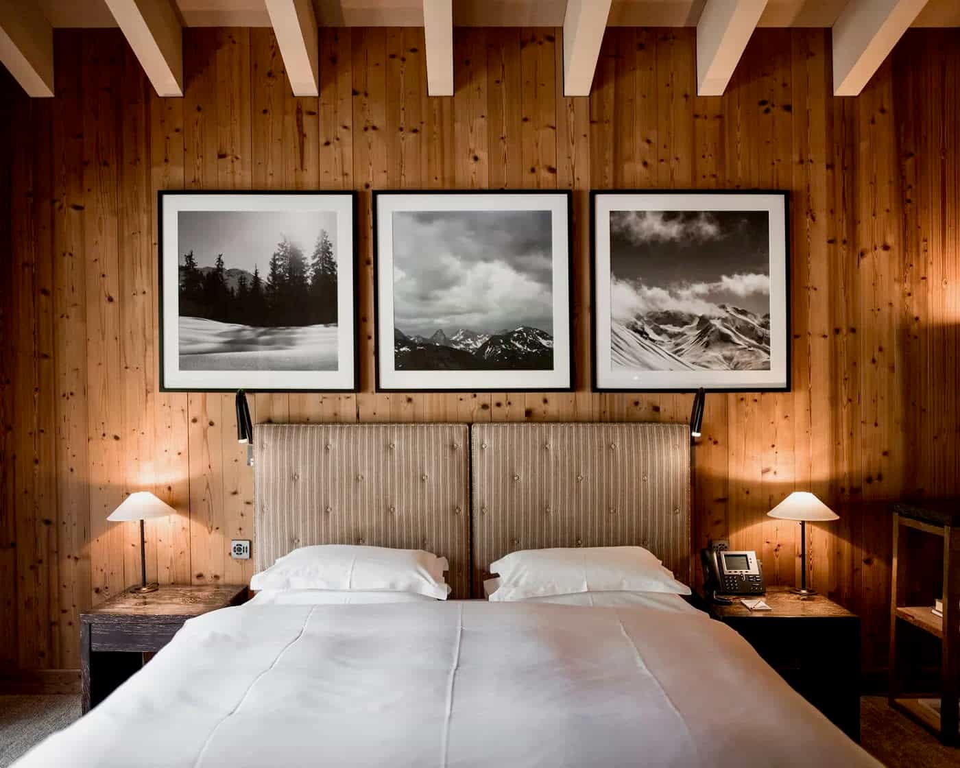 hotel room with white linen bed, wood wall and beam ceiling ith black and white framed photos above the bed