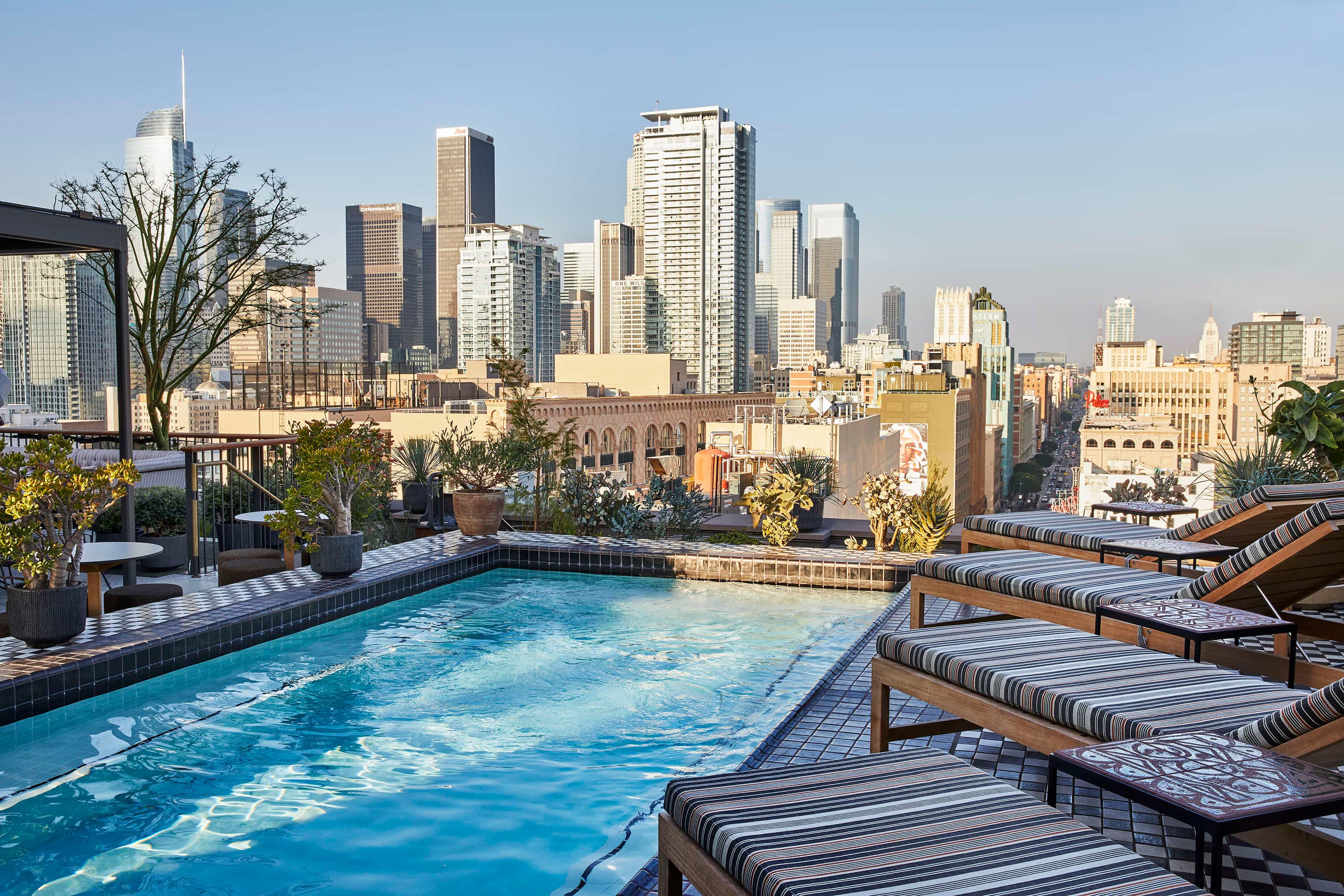 Rooftop pool with striped lounge chairs and potted plants overlooking the Los Angeles skyline under a clear afternoon sky.