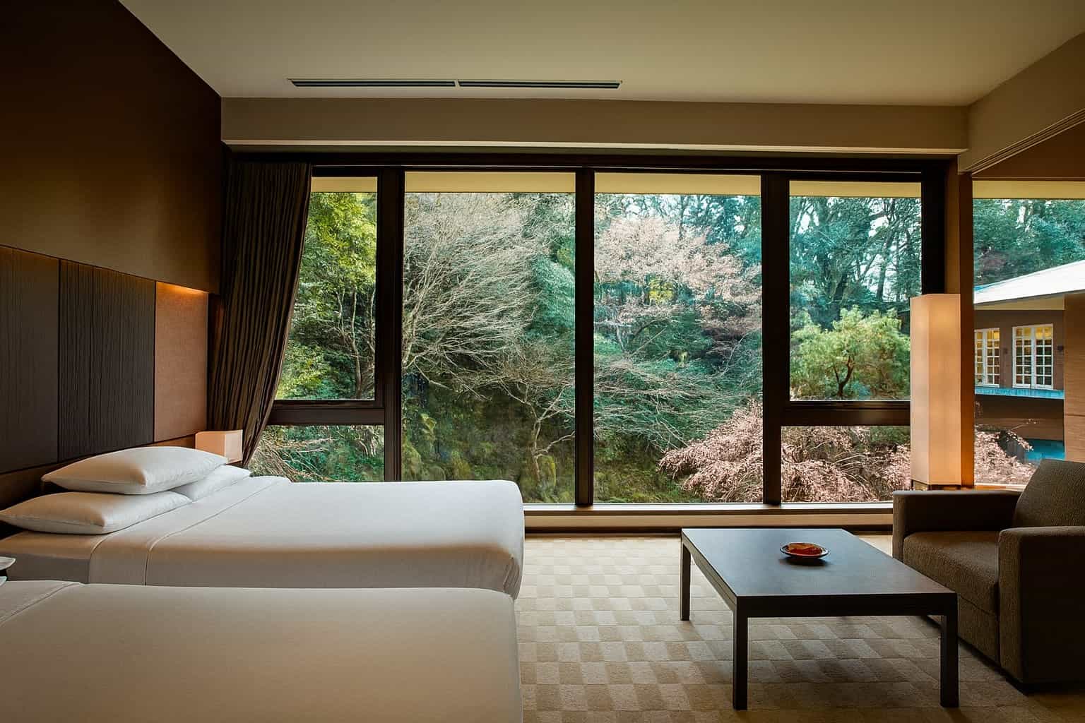 Spacious hotel room with two white beds facing a floor-to-ceiling window that overlooks a lush garden with trees and seasonal foliage.