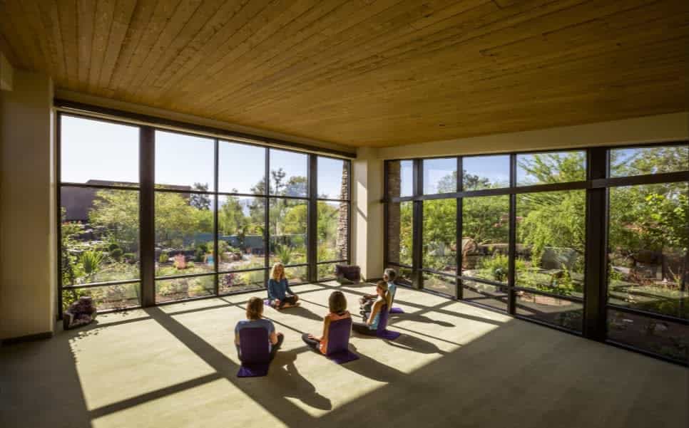 A spacious yoga studio with floor-to-ceiling windows overlooks a lush garden, as a small group of people sit on mats in a circle on the sunlit floor.