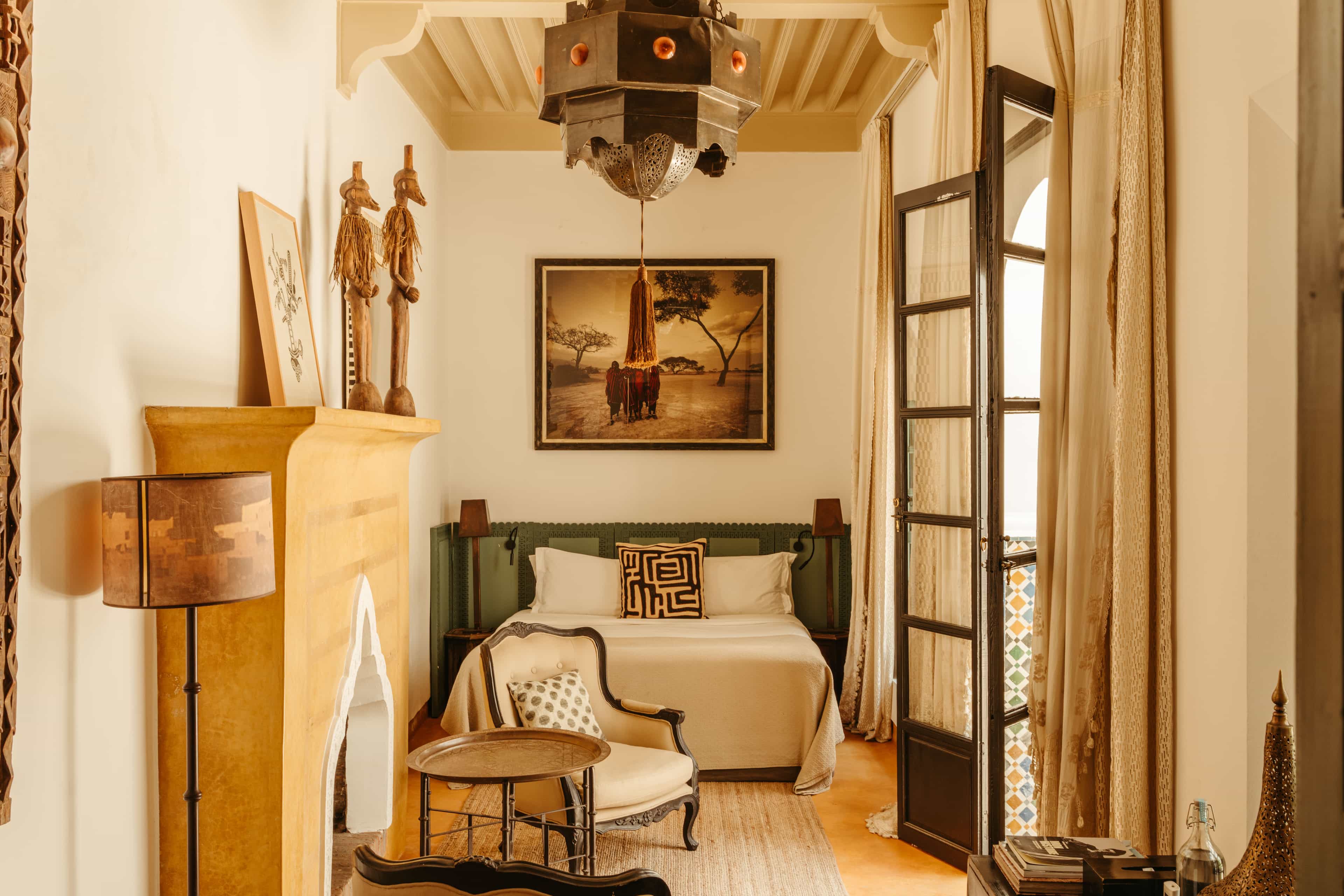 Warm-toned Moroccan bedroom featuring a carved plaster fireplace, brass lantern ceiling light, patterned textiles, and French doors opening onto a tiled balcony.