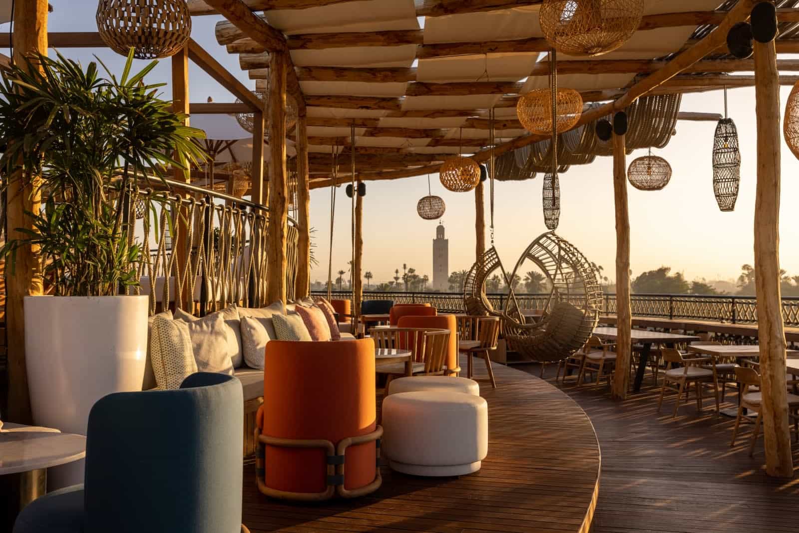 Rooftop terrace at Nobu Marrakech with woven lanterns, timber pergola beams, low lounge seating, and hanging rattan chairs, set against golden-hour views of the Medina and Koutoubia Mosque in the distance.