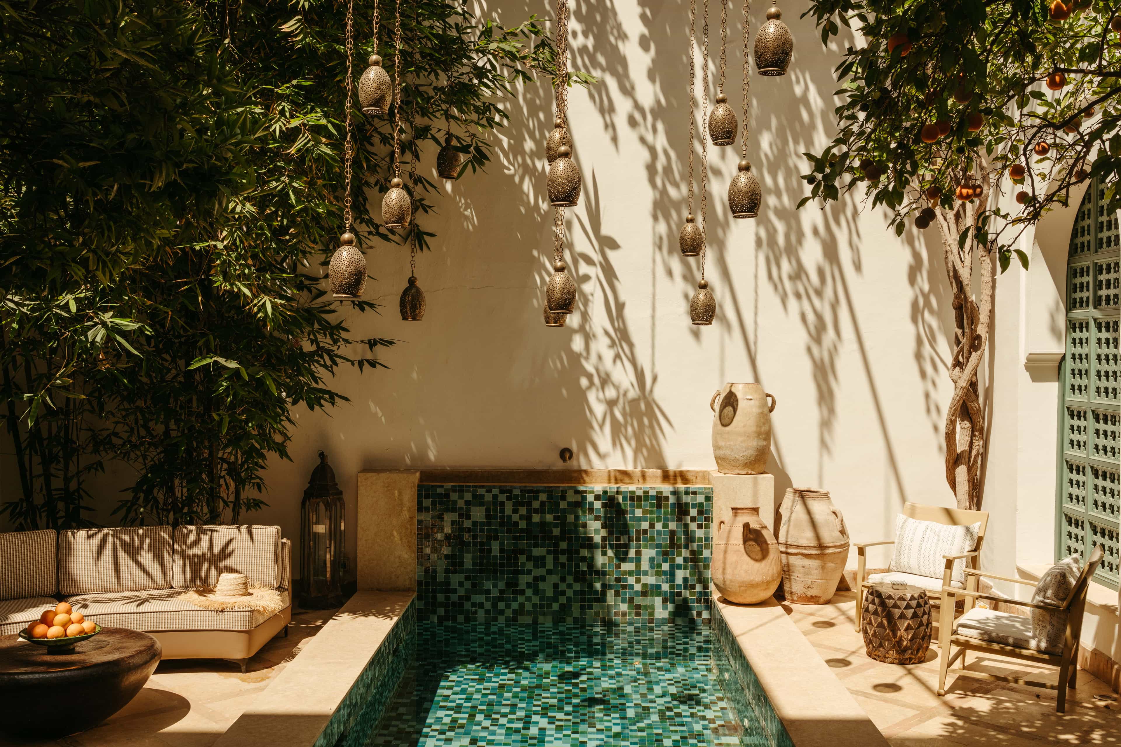A serene riad courtyard with a small mosaic-tiled plunge pool, hanging brass lanterns casting patterned shadows on cream walls, leafy trees overhead, and relaxed seating with pottery accents in warm, earthy tones.