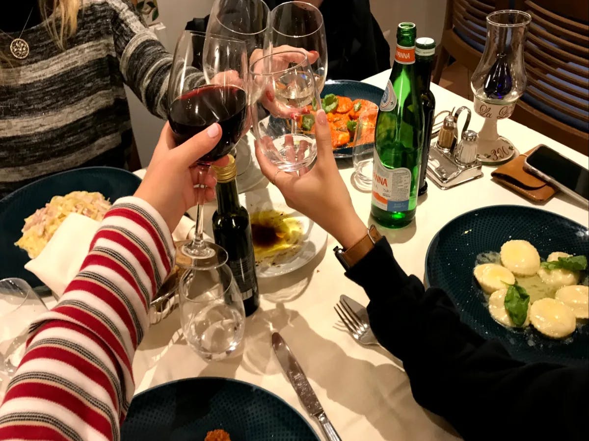A picture of three people holding a wine glass up and gesturing a cheers over a table of food and beverages.