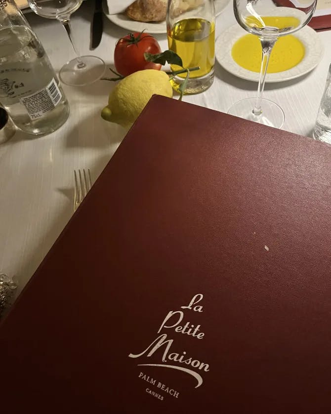 Photo of a red/brown menu for "Le Petit Maison" on a table with a wine glass.
