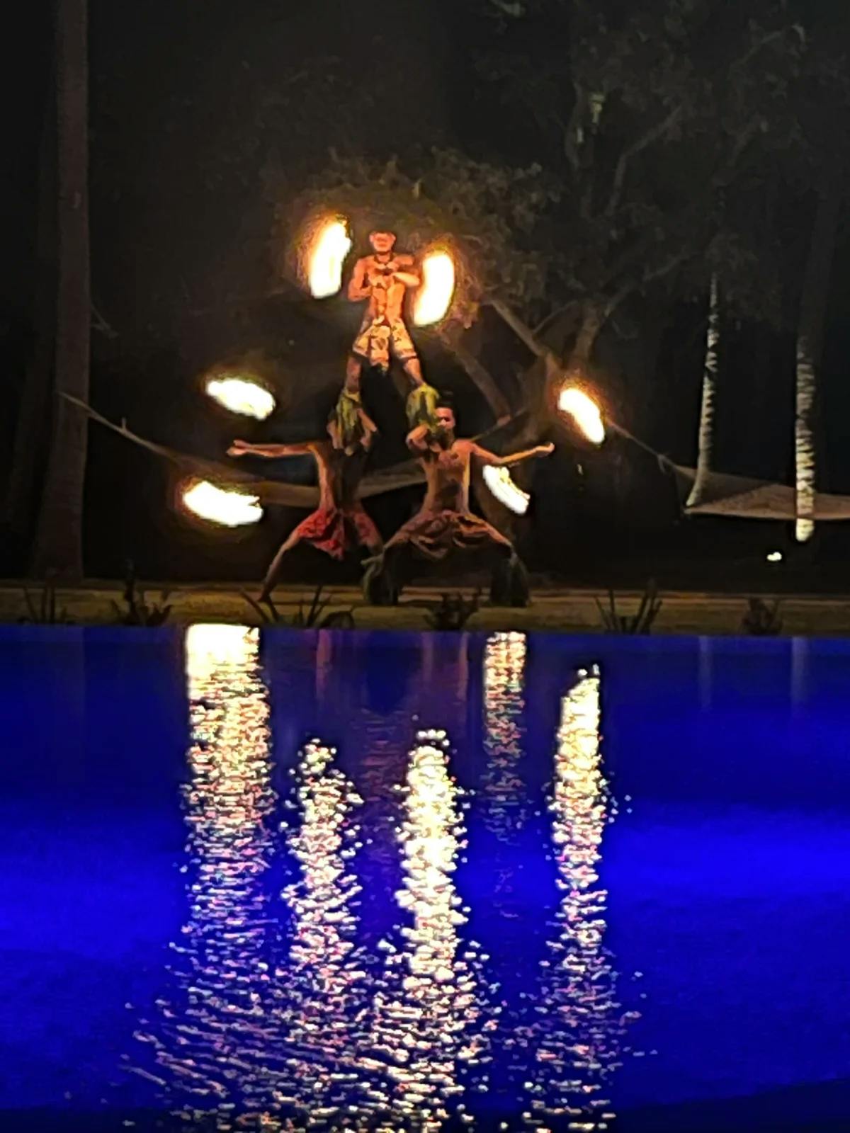 Three men in sarongs standing in a human pyramid holding flaming torches next to the pool, during the weekly fire dance dinner show