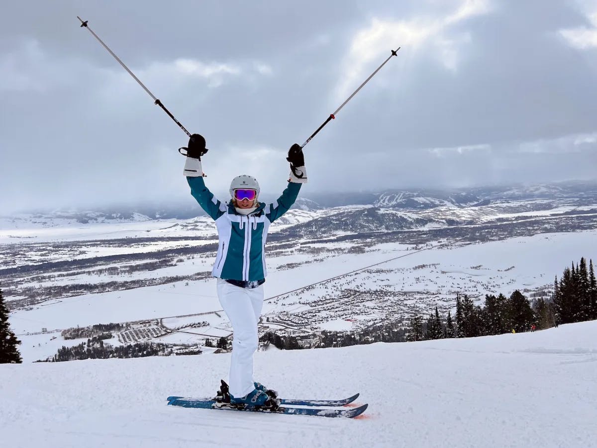 A person outside wearing ski gear on top of a slope with their arms and ski poles outstretched above their head. There is a view of snowy terrain and mountains in the cloudy distance behind them. 