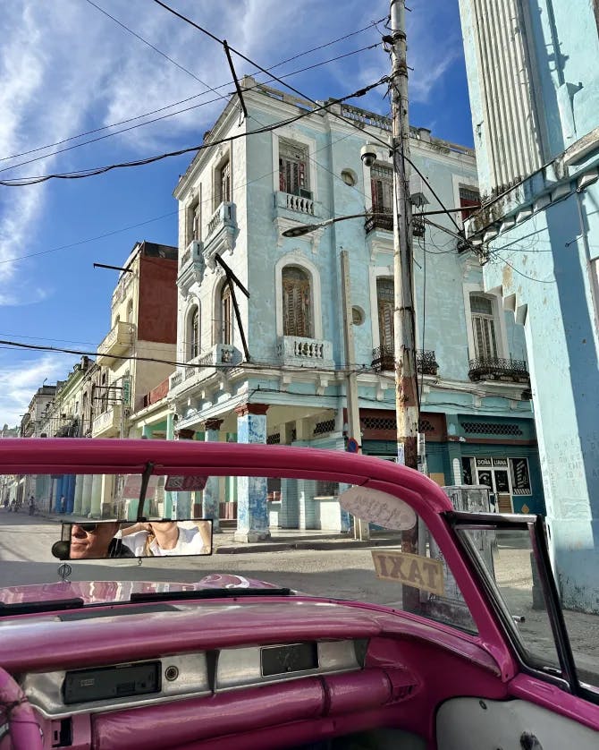 Light blue buildings seen from the inside of a vintage, pink convertible car. 