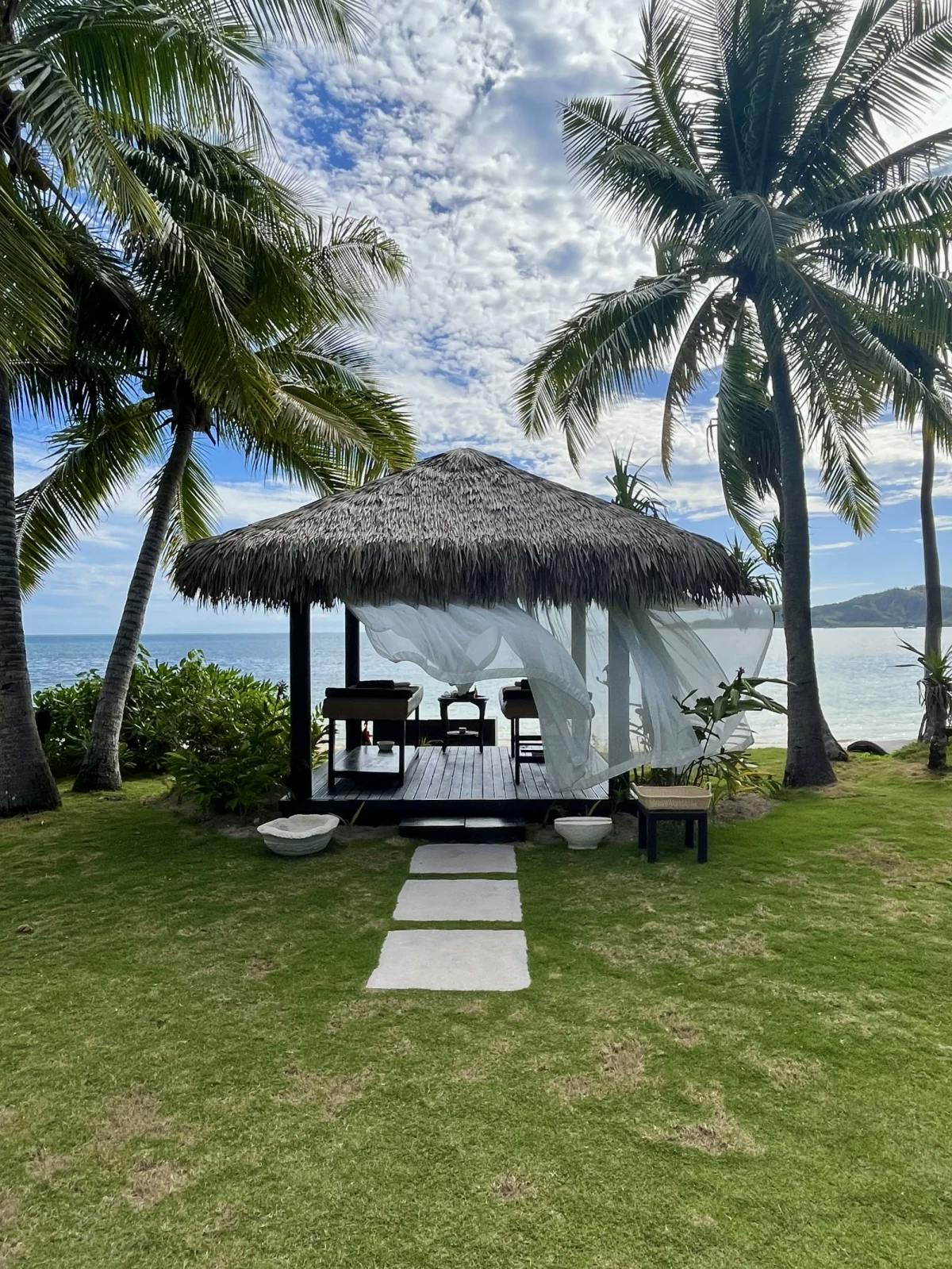 A private beachfront massage bure, a thatched hut with gauzy curtains overlooking the ocean, with palm trees on either side