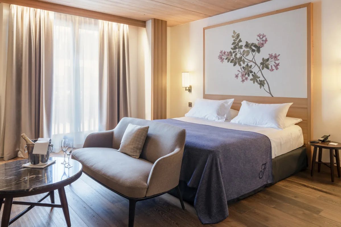 A room at the Faloria Mountain Spa Resort, with a bed with blue comforter, couch, wood-paneled ceiling, botanical painting and a table set with champagne.