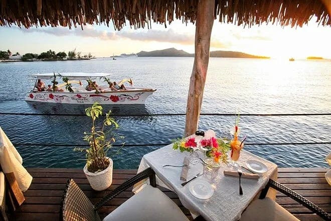A beautifully set table with flowers and drinks, on the balcony of a an overwater bungalow, with a view of the sea, a boat, mountains and the soon-to-be-setting sun.