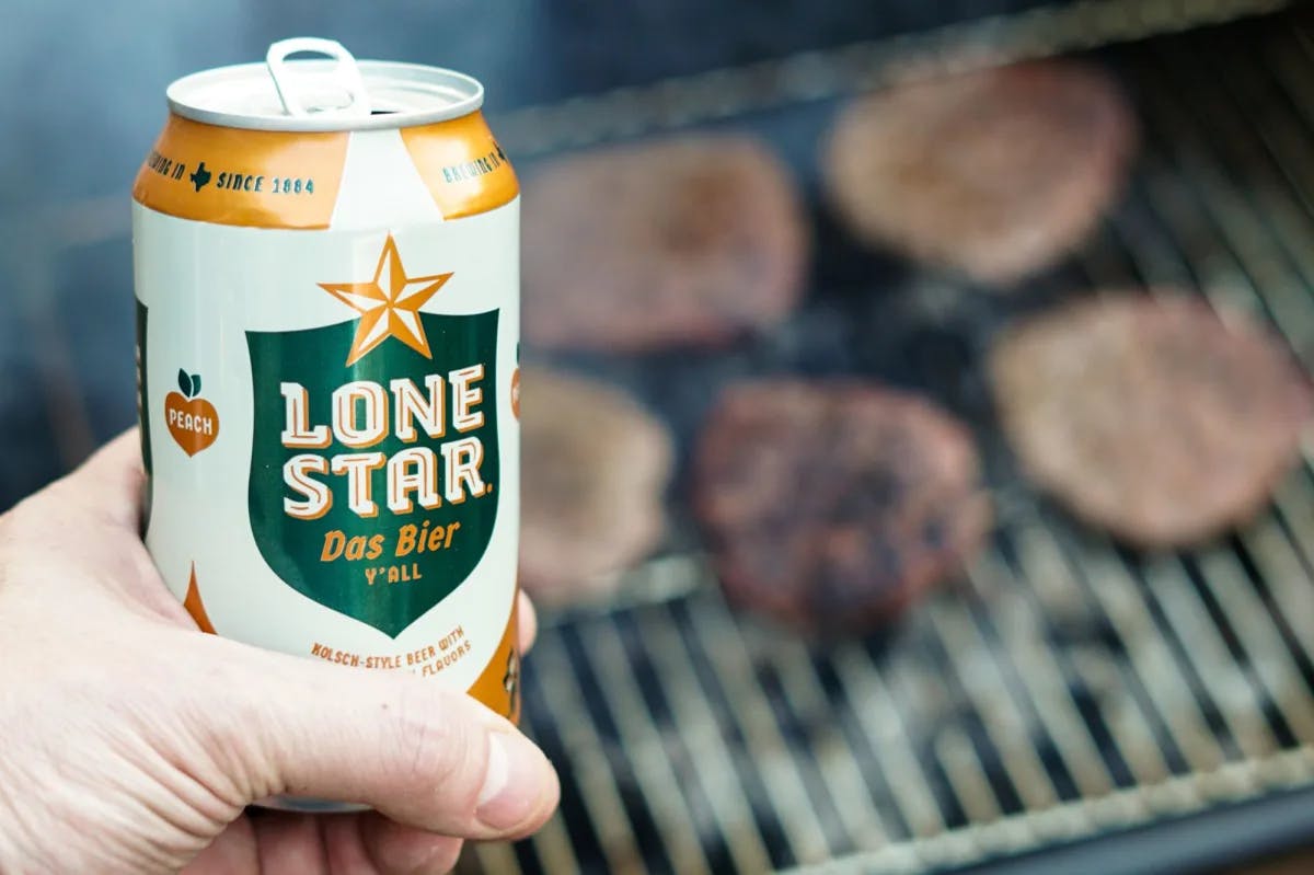 A person holding Lone Star Can with BBQ on grill at the back.