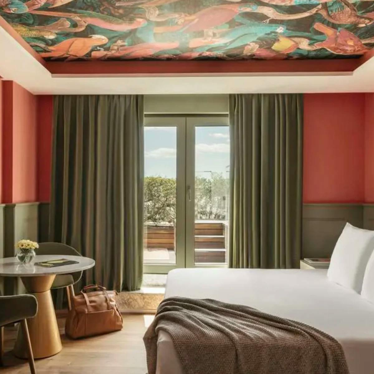 A bedroom in the Hotel Colon, a Gran Melia hotel, with red and green walls, an abstract mural on the ceiling and a view of trees from the balcony.