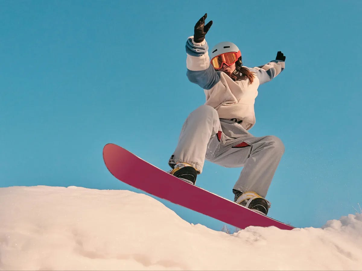 A snowboarder in white gear executes a mid-air trick against a backdrop of clear blue skies and snowy terrain.