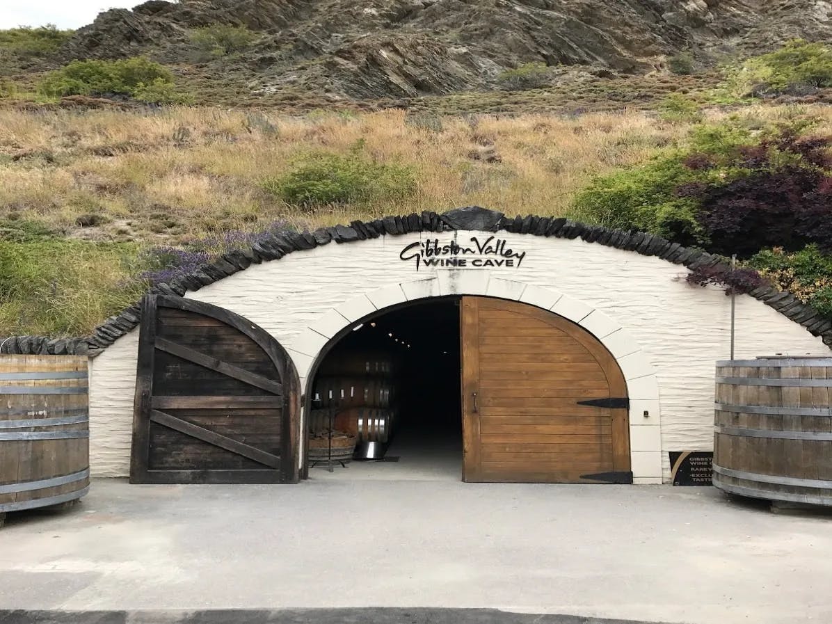 A picture of the entrance of a winery complete with a white archway nestled into a mountain with a wooden gate during daytime.