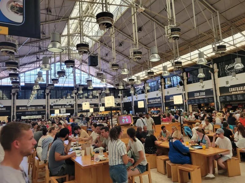 A food mall with people sitting on brown chairs and tables eating food. 