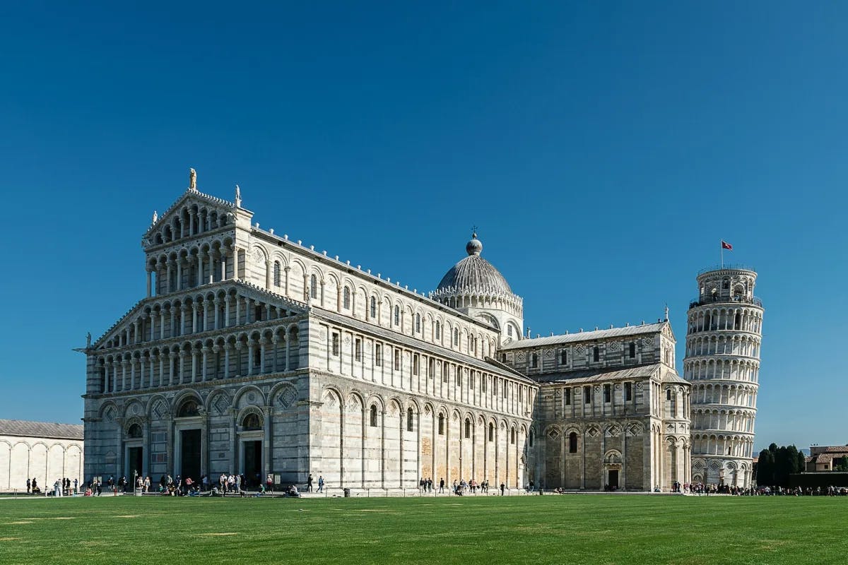A large, stone building with intricately detailed architecture on top of a green lawn and the leaning tower of Pisa to the right. 