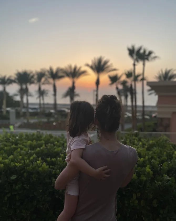 A photo of Jennifer holding her daughter and looking out toward palm trees, bushes and an orange sunset. 