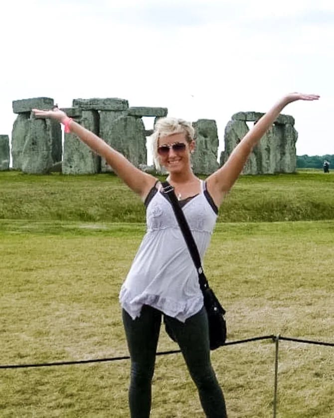 A woman stands with her arms in the air in front of Stonehenge.