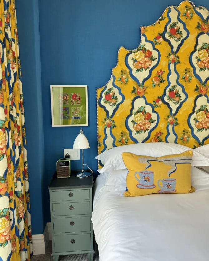 A hotel suite with a colorfully patterned headboard, white bedding, blue wall and teal nightstand with a lamp sitting on top of it and piece of artwork hanging behind it. 
