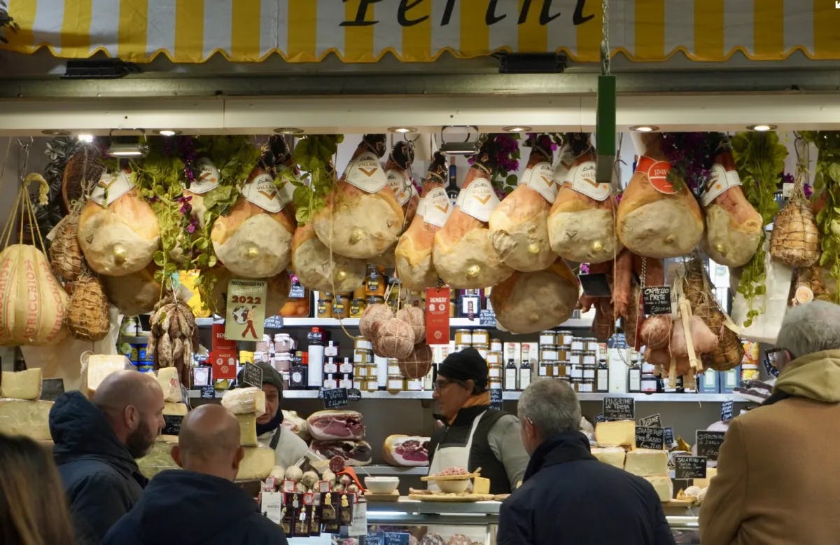 A view of a market with cured meat hanging, people shopping and a yellow and white striped awning. 