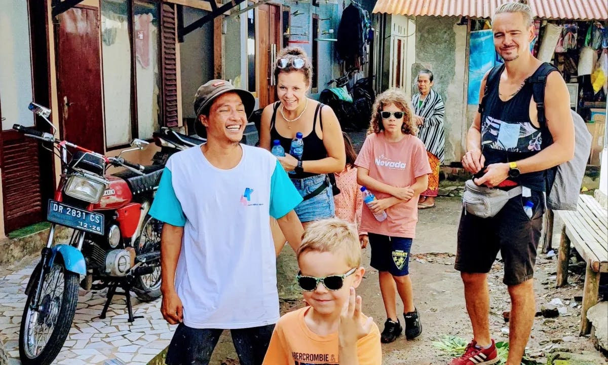 This image depicts a group of people gathered around outside and smiling. There is Indonesian architecture and a motorized bike in the surrounding area. 
