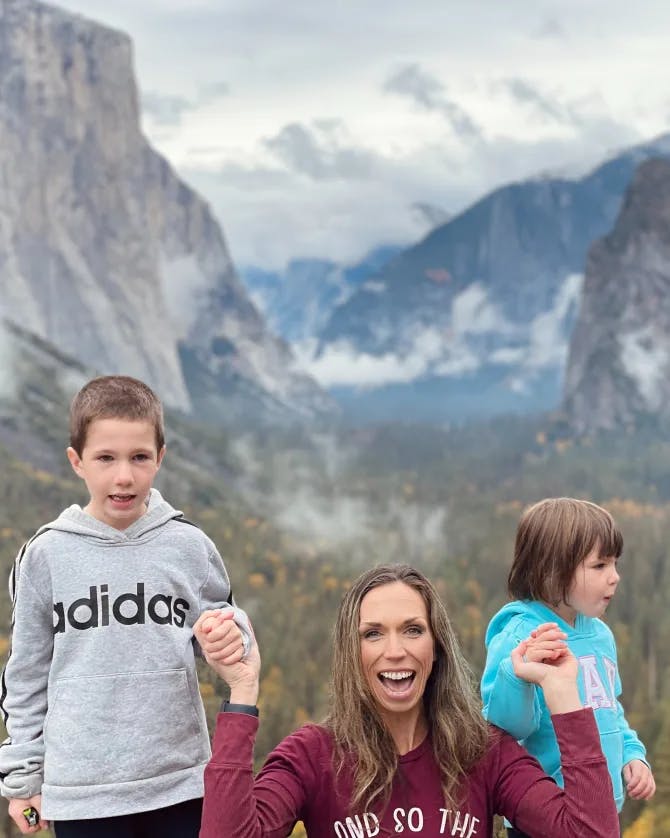 A photo of Jennifer smiling and holding her childrens hands in front of a forest and large mountains.