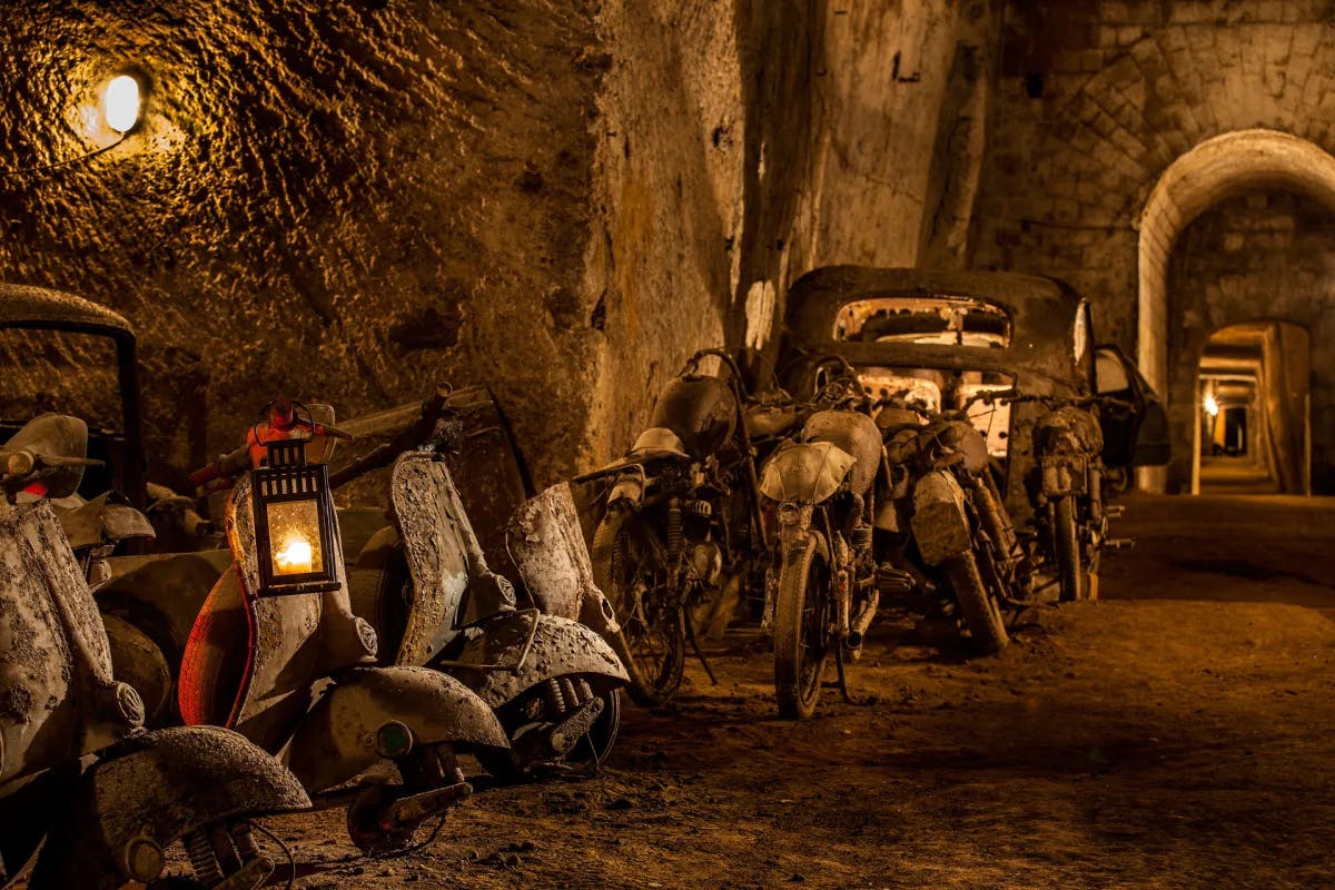 A view of a rocky tunnel with old vespas, lanterns and cars leading towards an archway. 