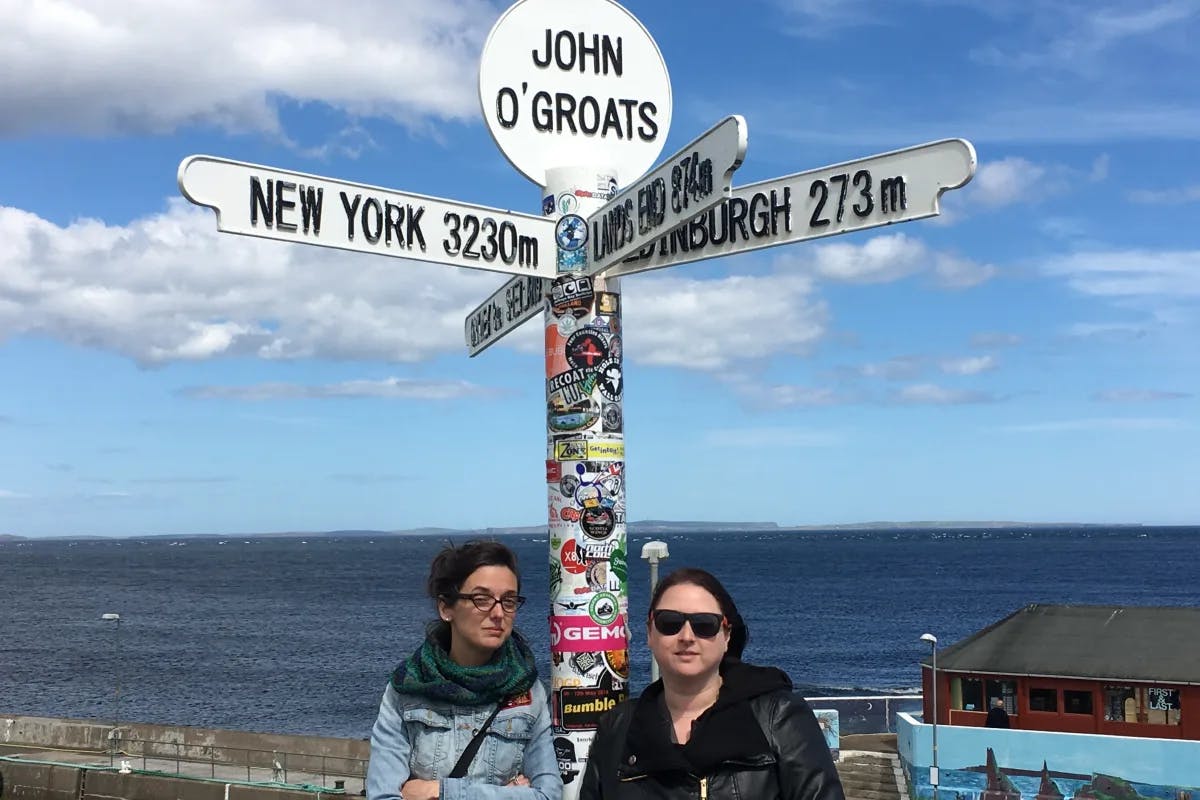 Two people posing under a white street sign covered in stickers that reads "John O'Groats" at the top. There is a blue sea in the background. 